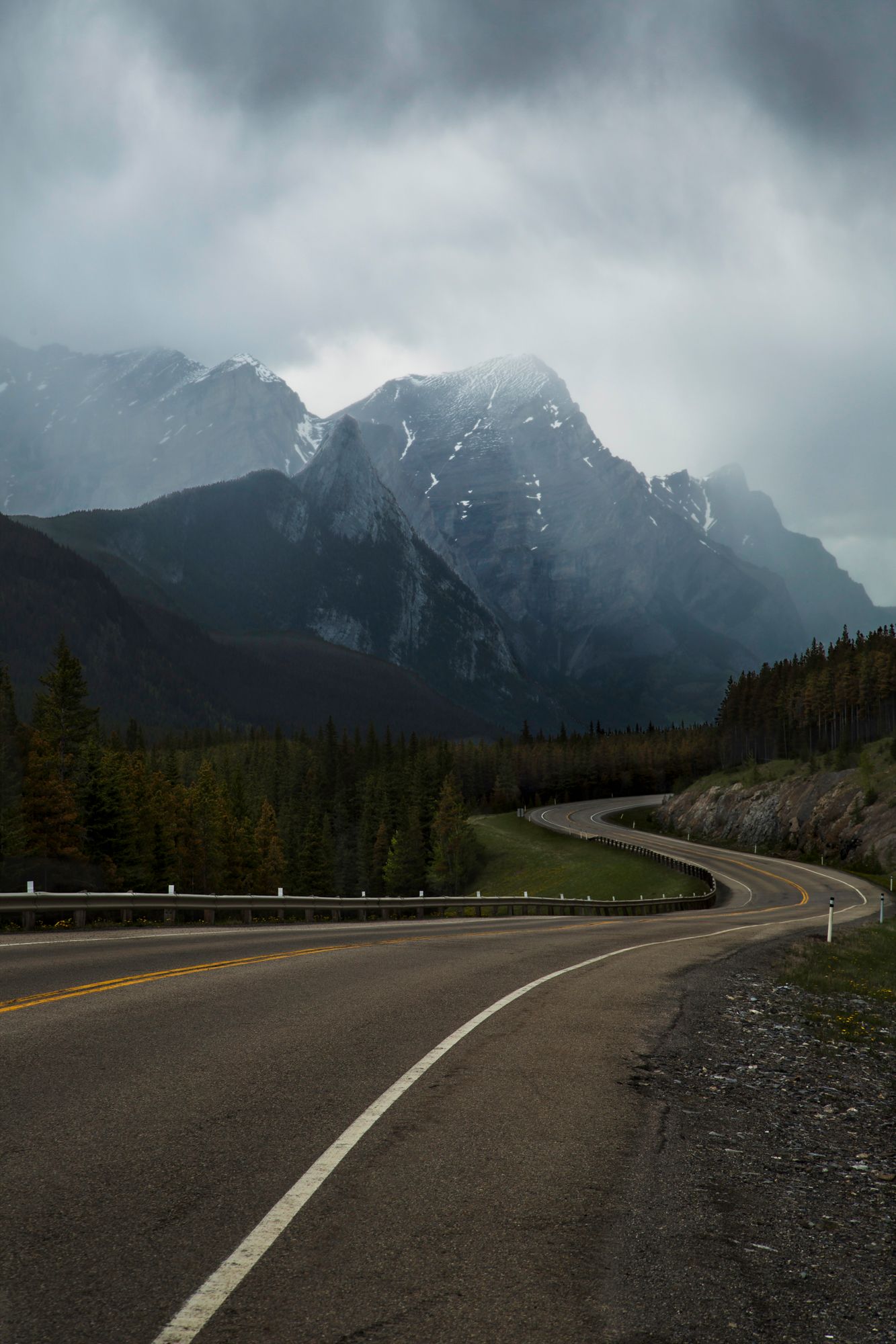 Winding mountain road curves through misty Canadian Rockies with snow-capped peaks and evergreen forest.