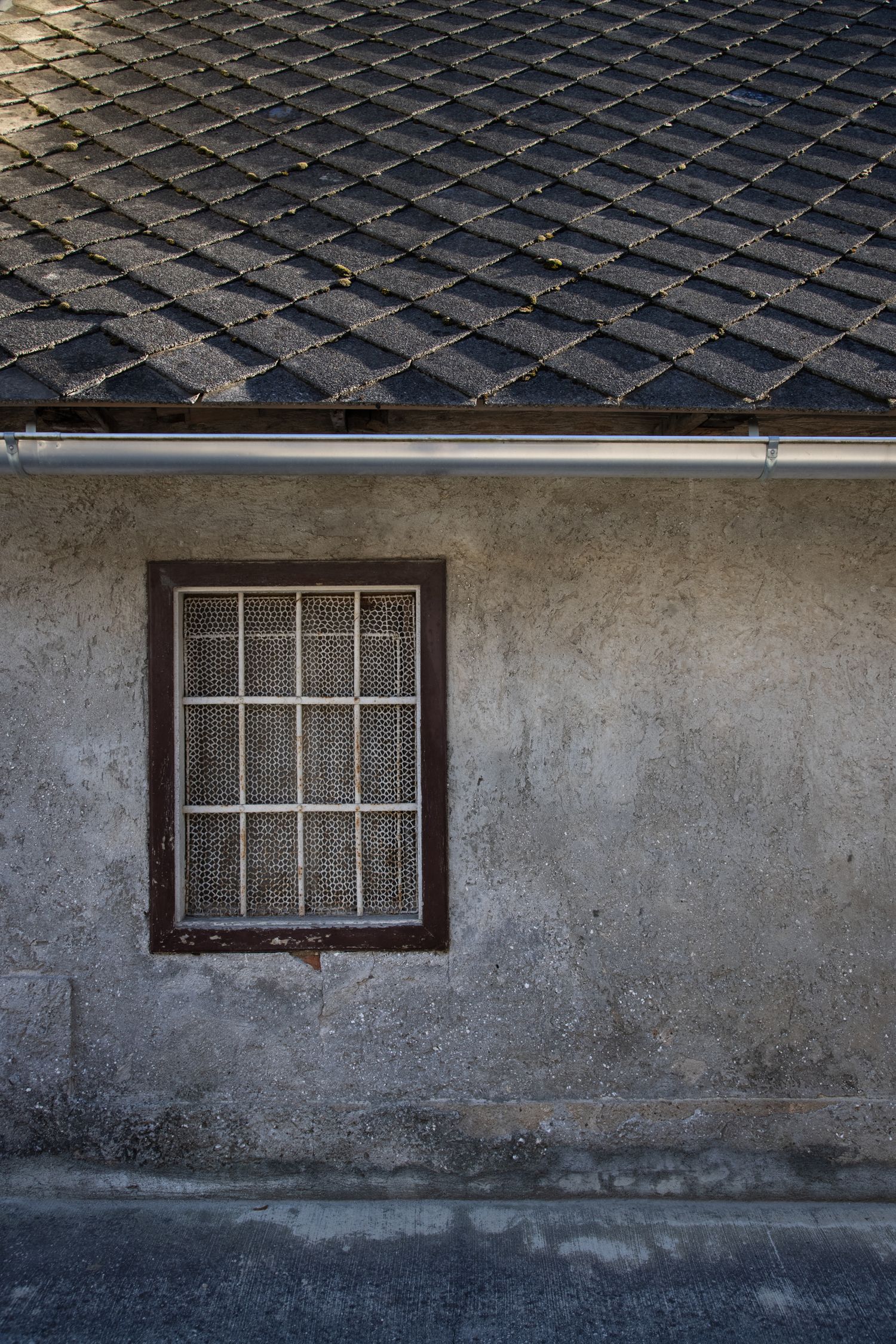 Old weathered building exterior with barred window, gray stucco wall, metal drainpipe, and dark diamond-patterned roof tiles.
