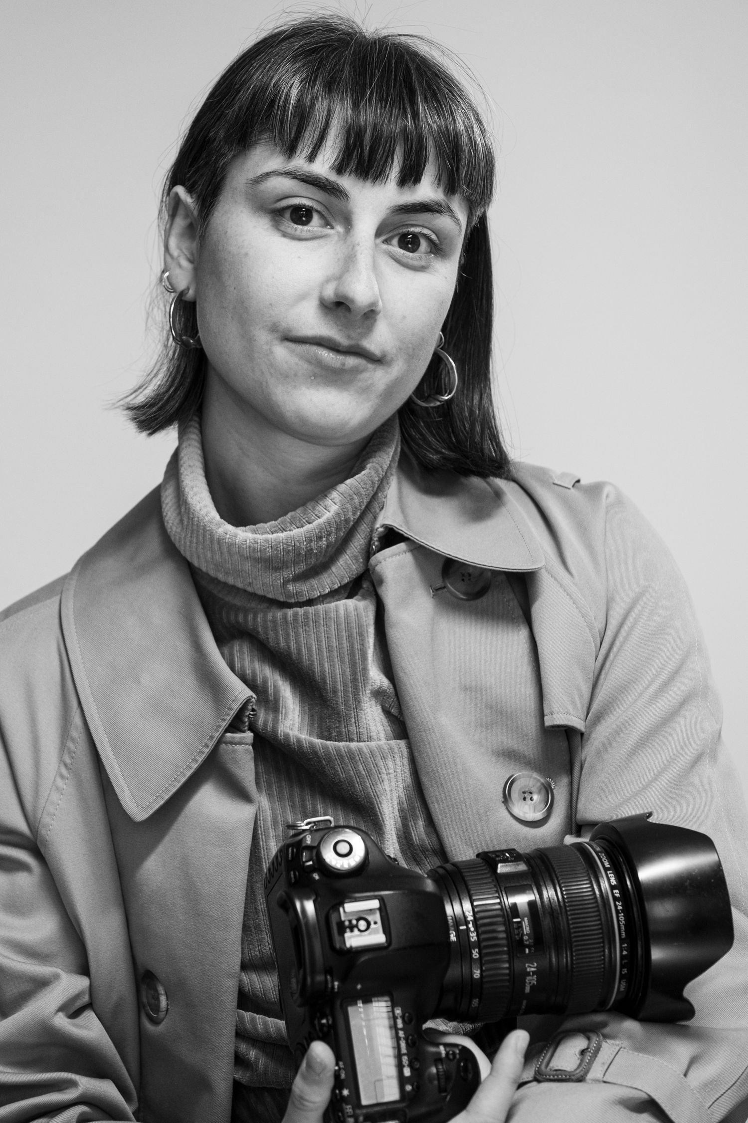 Black and white portrait of a photographer wearing a turtleneck and trench coat while holding a professional camera.