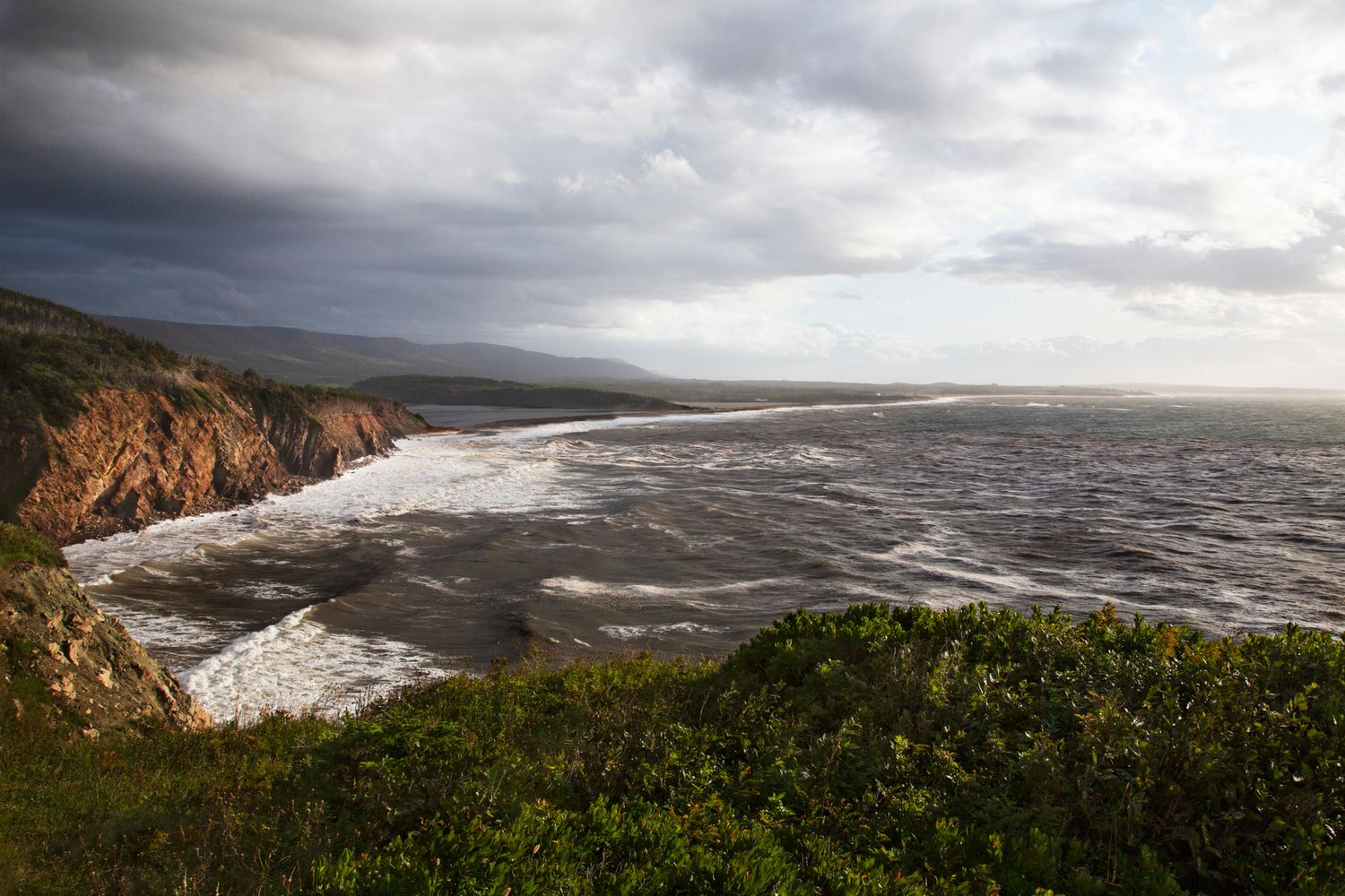 Sweeping view of rugged coastline with dramatic cliffs and churning ocean under stormy skies.