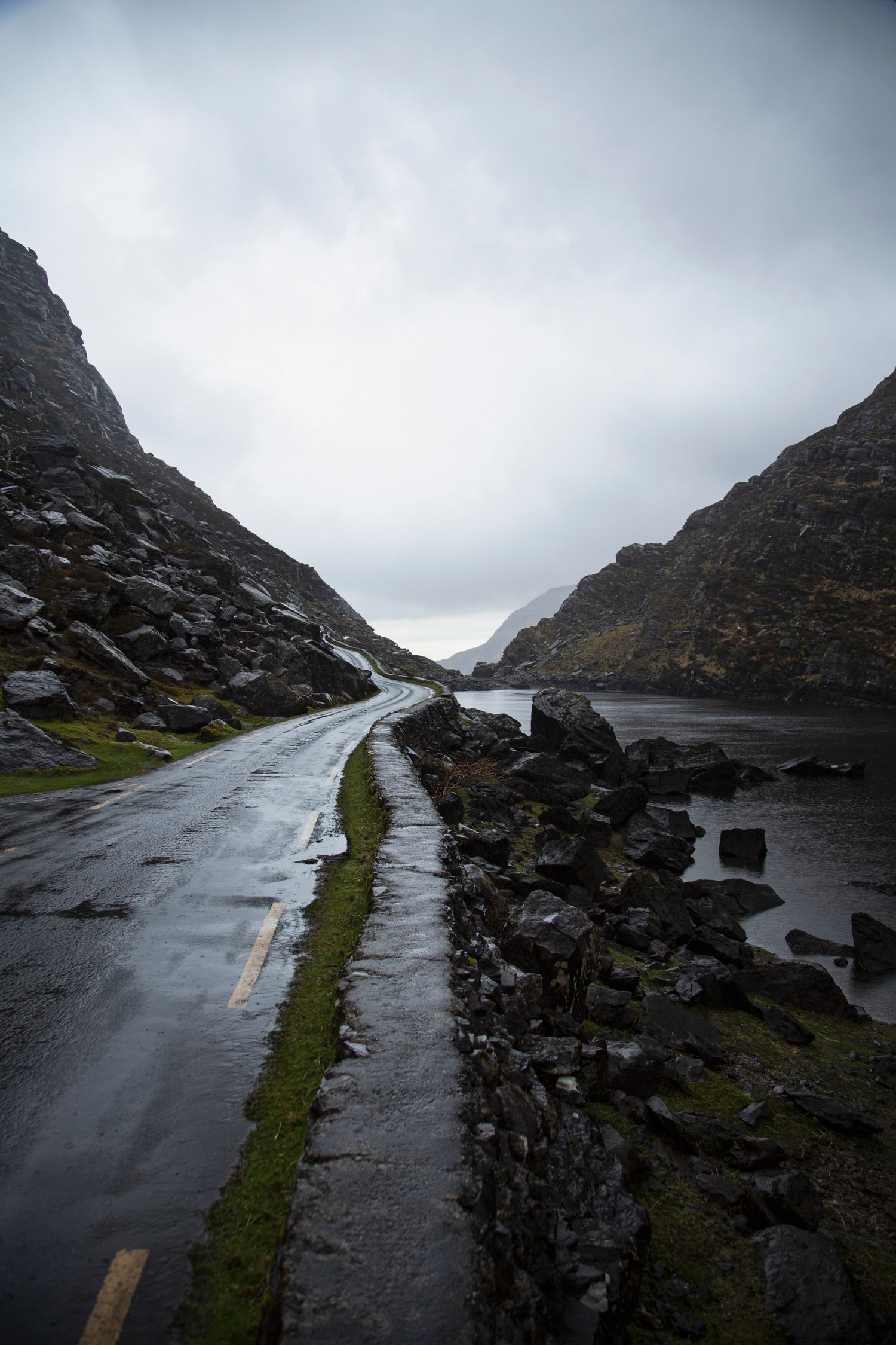 Wet mountain road curves through rocky landscape with grass verges on a gloomy day.