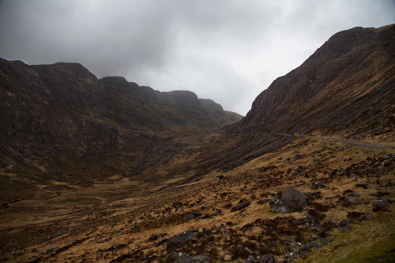 Panoramic view of rugged Scottish Highlands with dramatic cloudy skies and winding mountain paths through the valley.