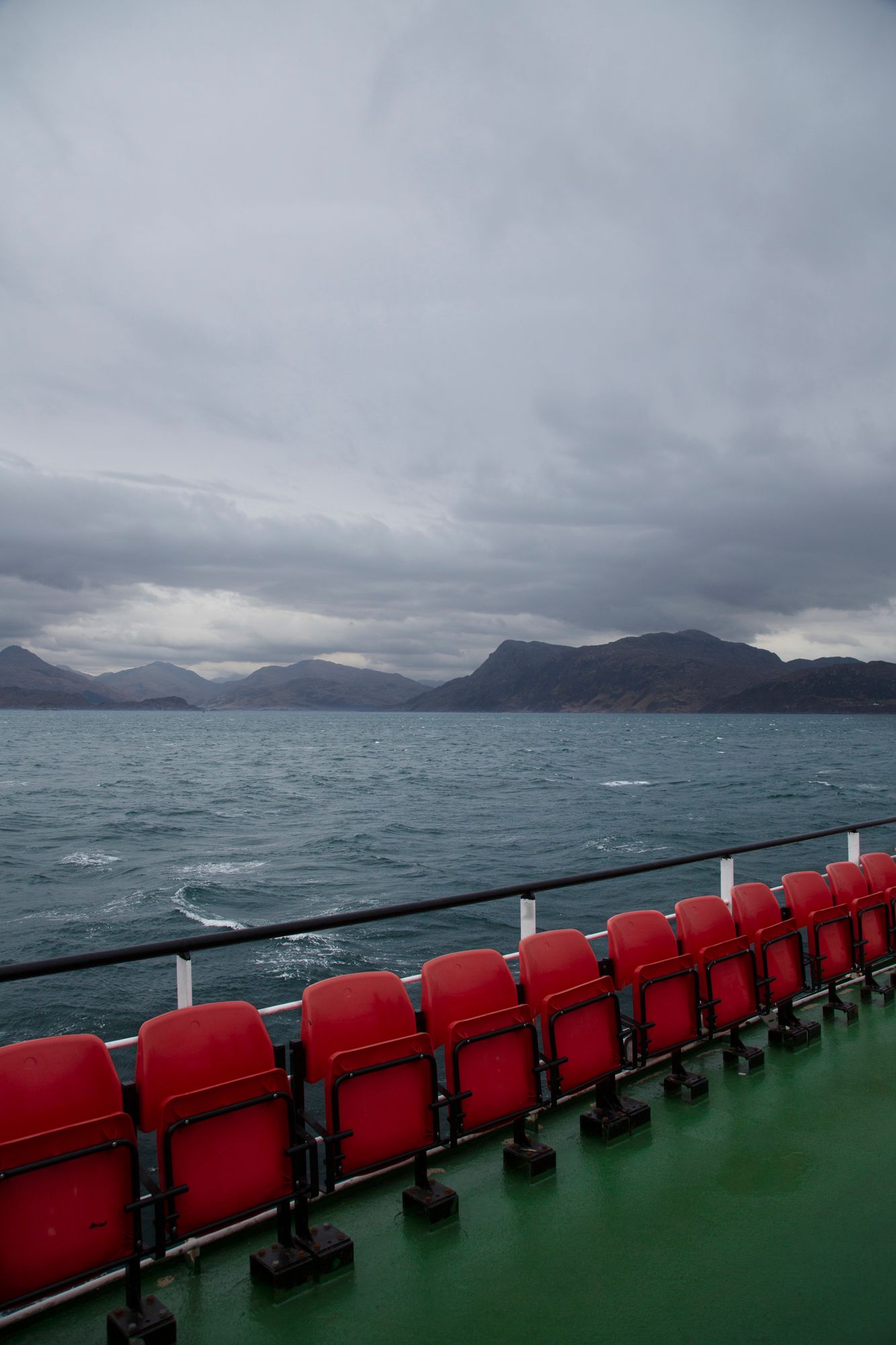 Red stadium seats line the deck of a ferry boat crossing choppy waters with misty mountains in the distance.