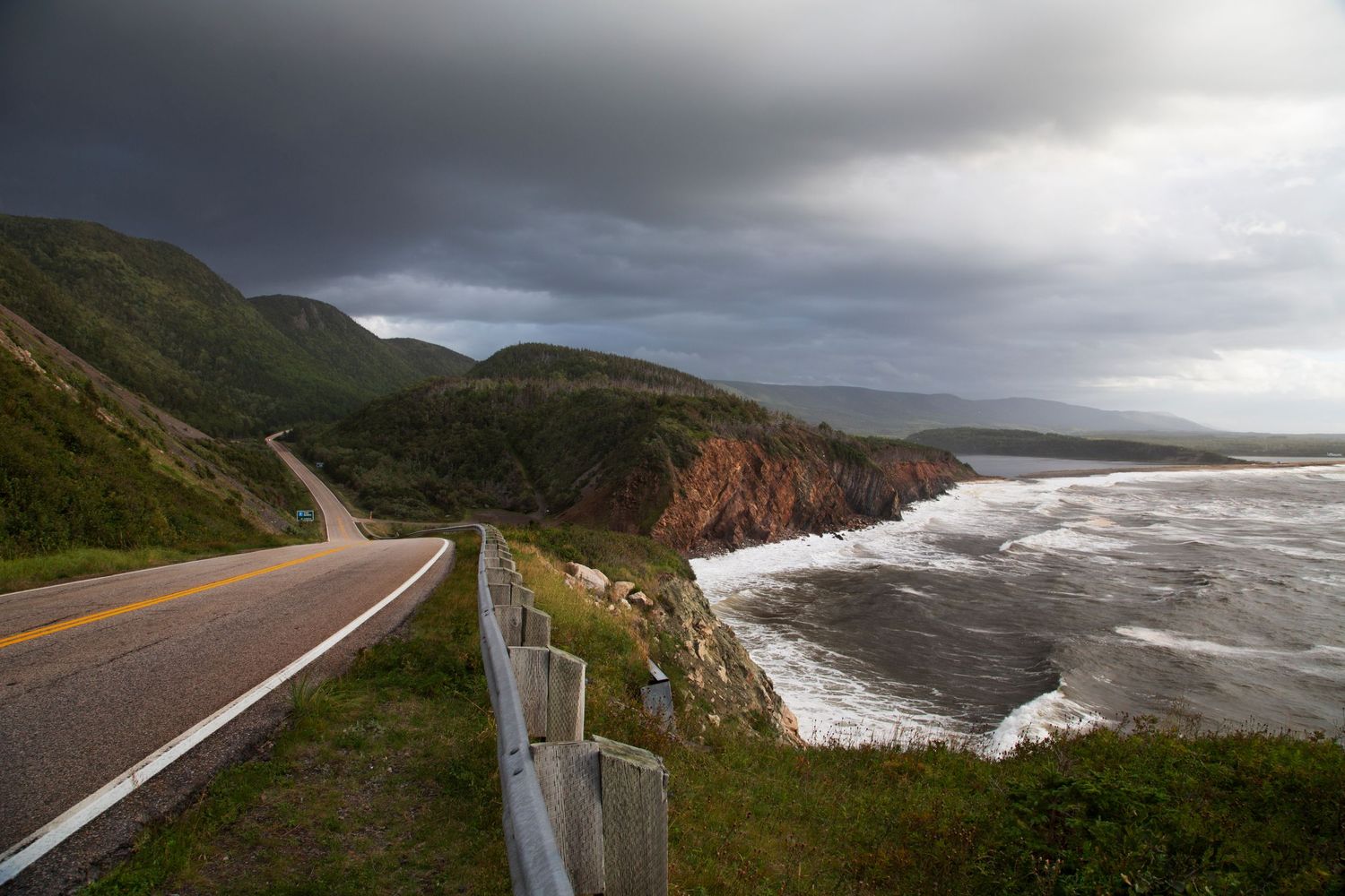 Dramatic coastal highway winding along cliffs with stormy skies and crashing waves below.