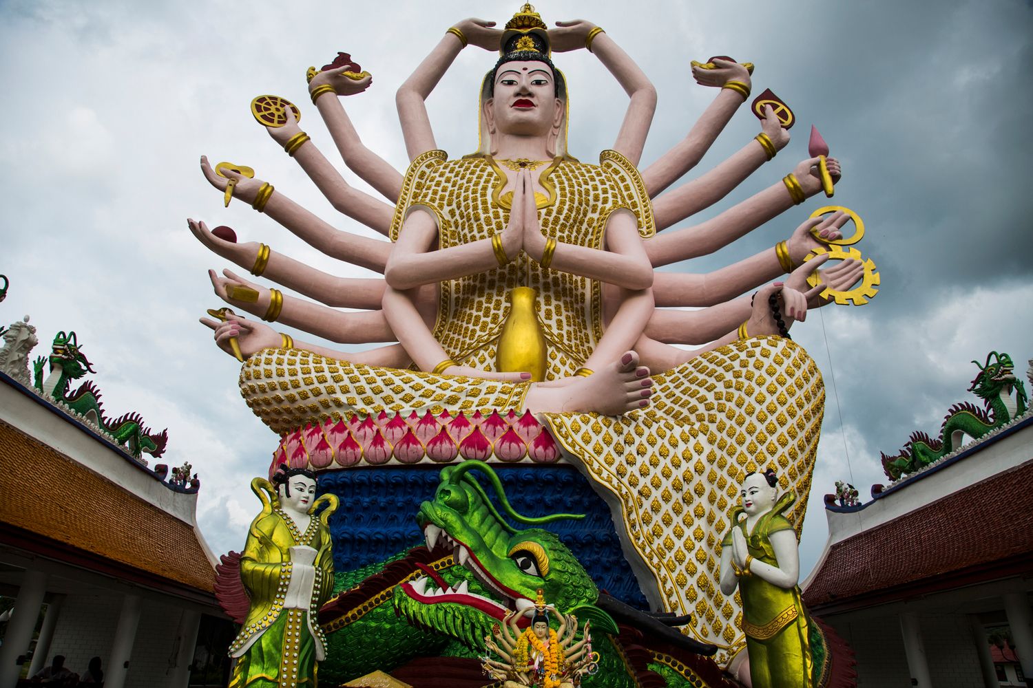 Multi-armed goddess statue in golden attire at a Buddhist temple complex.