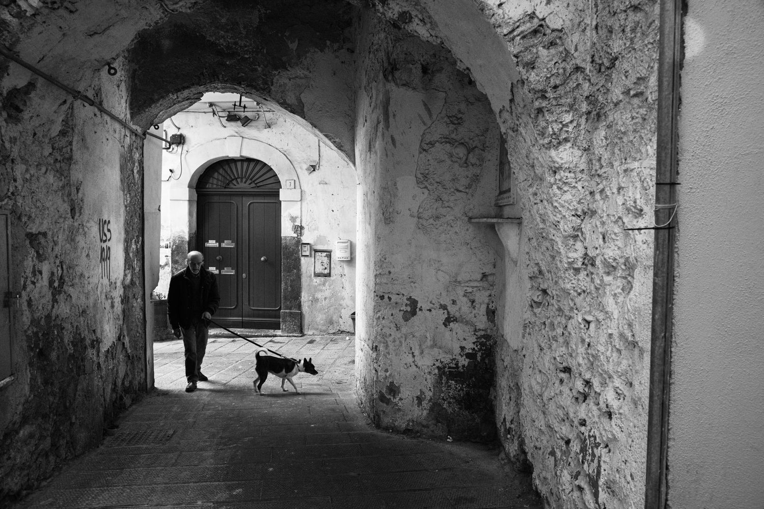 Black and white photo of a person walking a dog through an ancient stone archway in a historic European village.