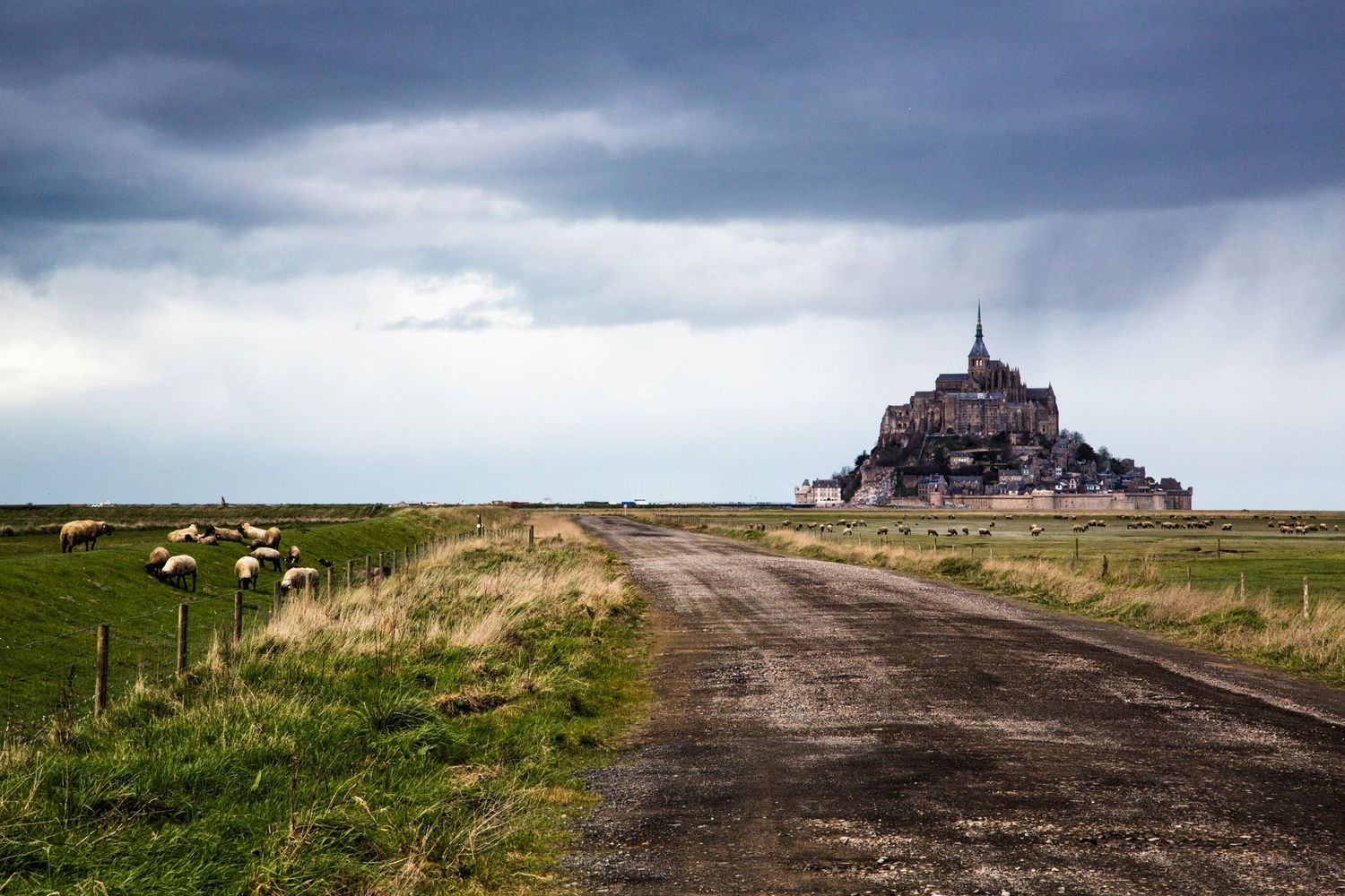 Long rural dirt road leading to a distant church under dramatic storm clouds.