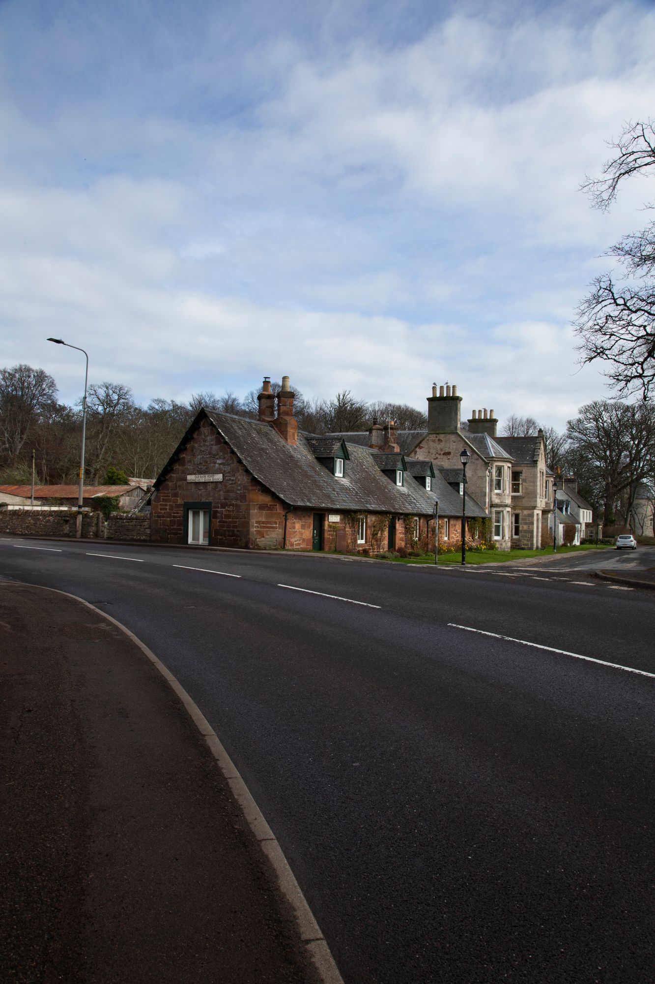 A curved rural road in Scotland with stone houses and buildings along the roadside under an overcast sky.