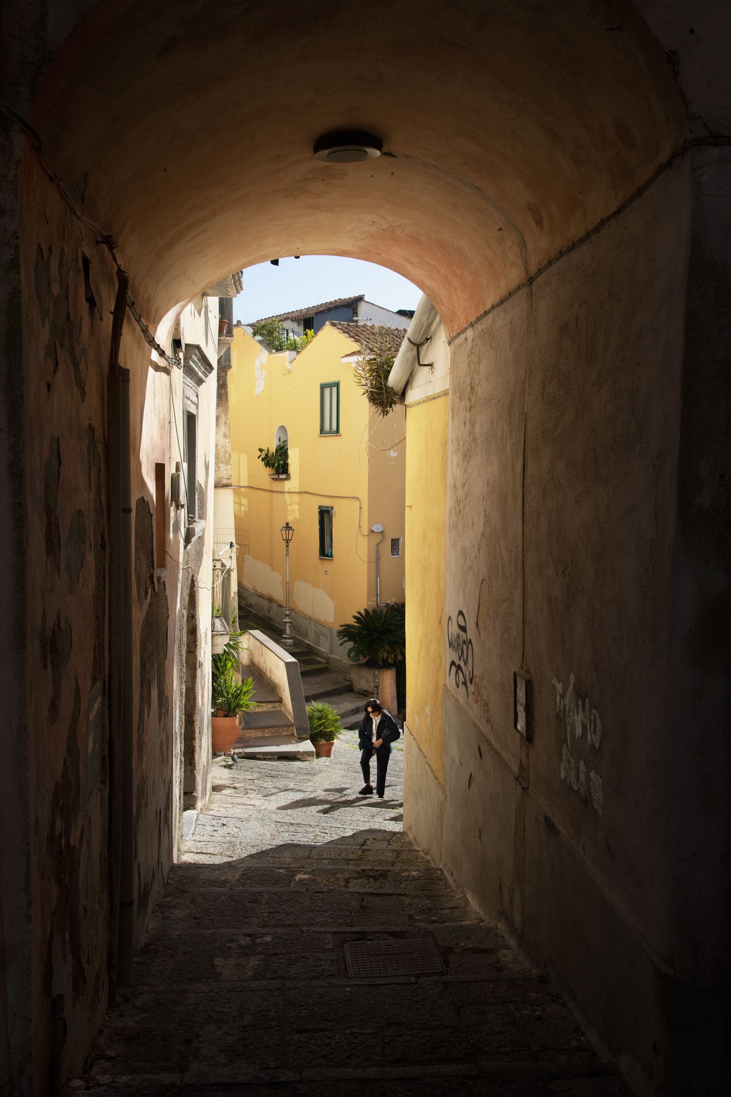 Person walking through a narrow arched alleyway in an Italian village with yellow buildings and stone steps.