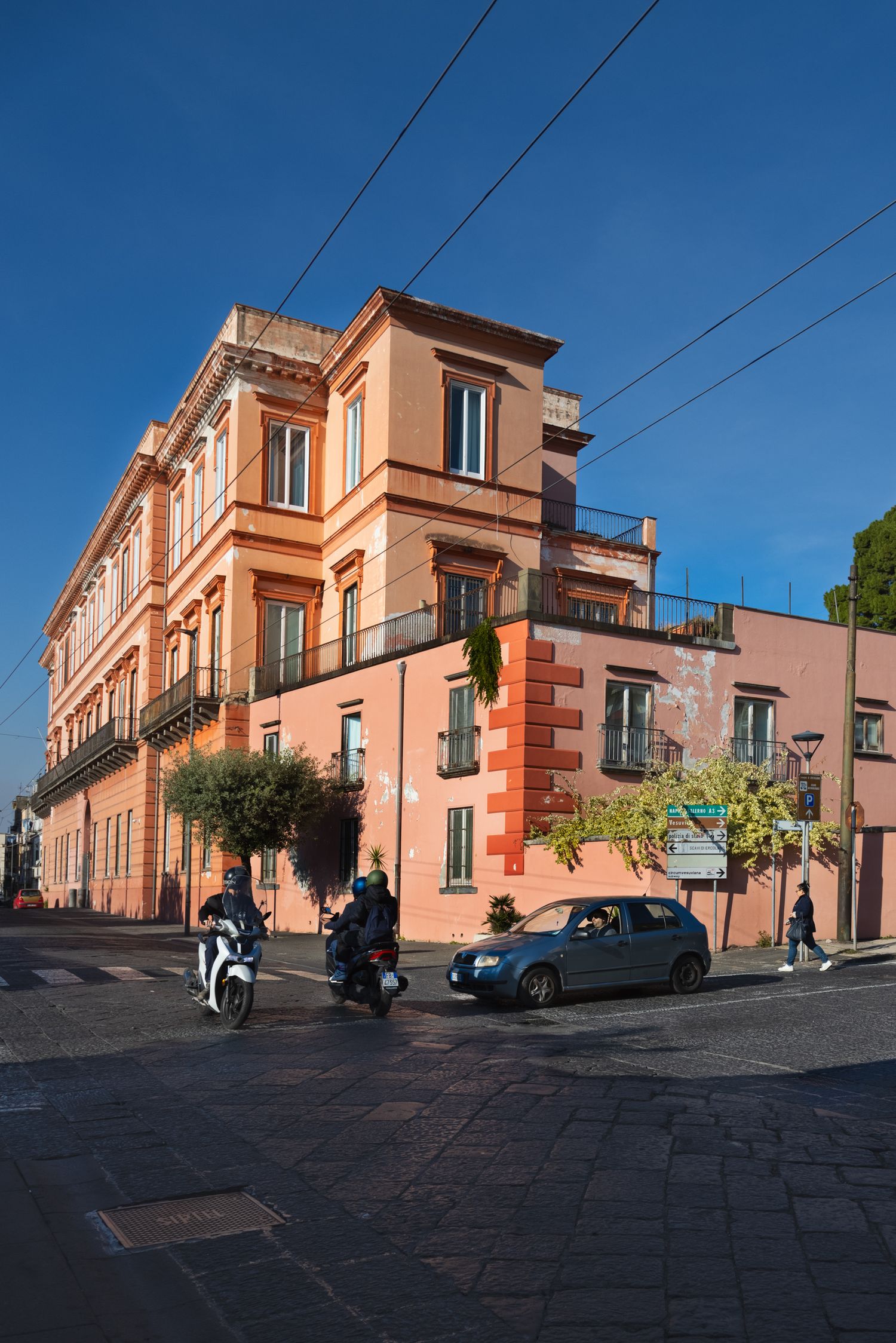 Motorcycles and car on a cobblestone street corner beside a terracotta Italian building under a clear blue sky.