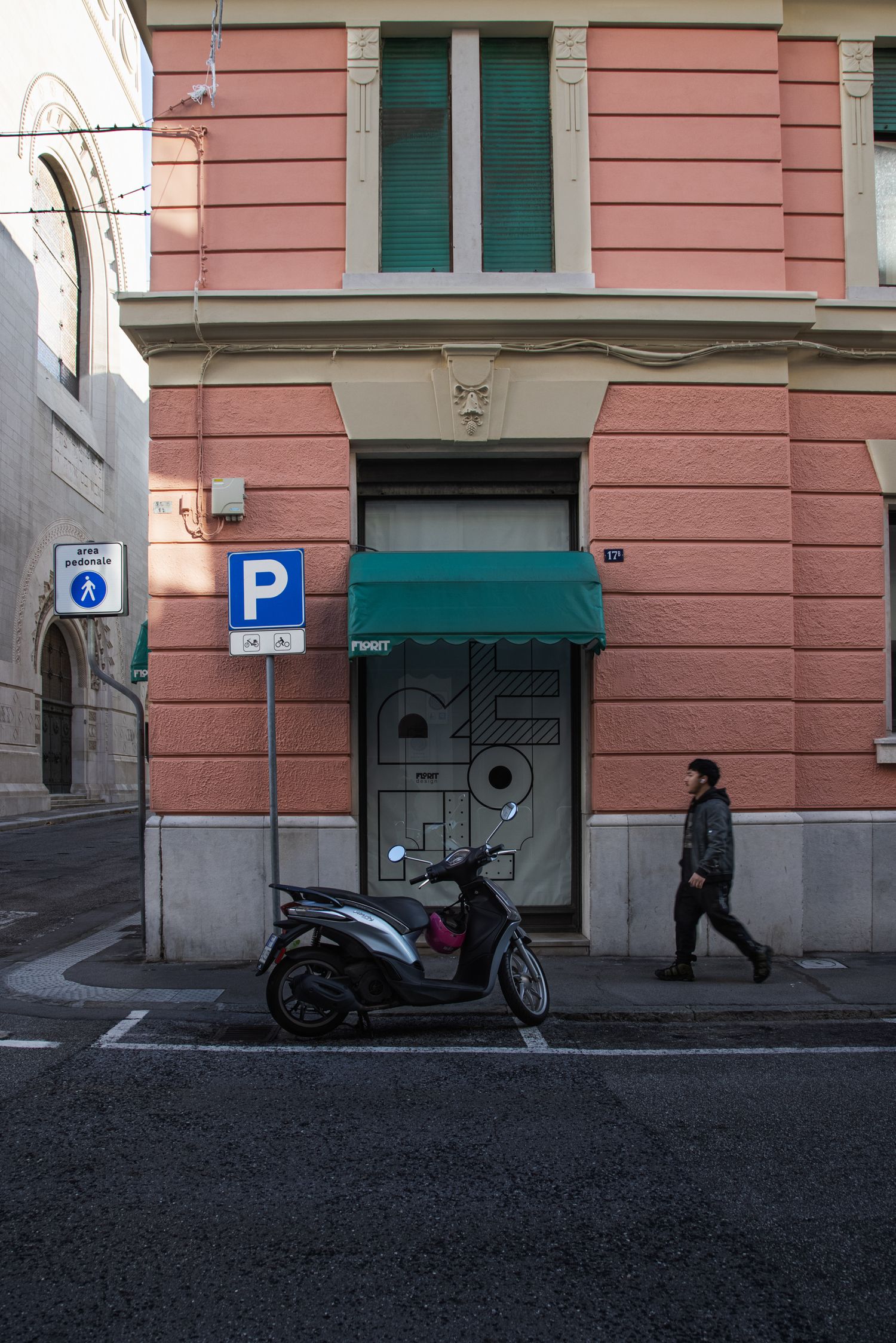 A scooter parked beside a blue parking sign on a sunlit European street corner with a pink building and green awning.