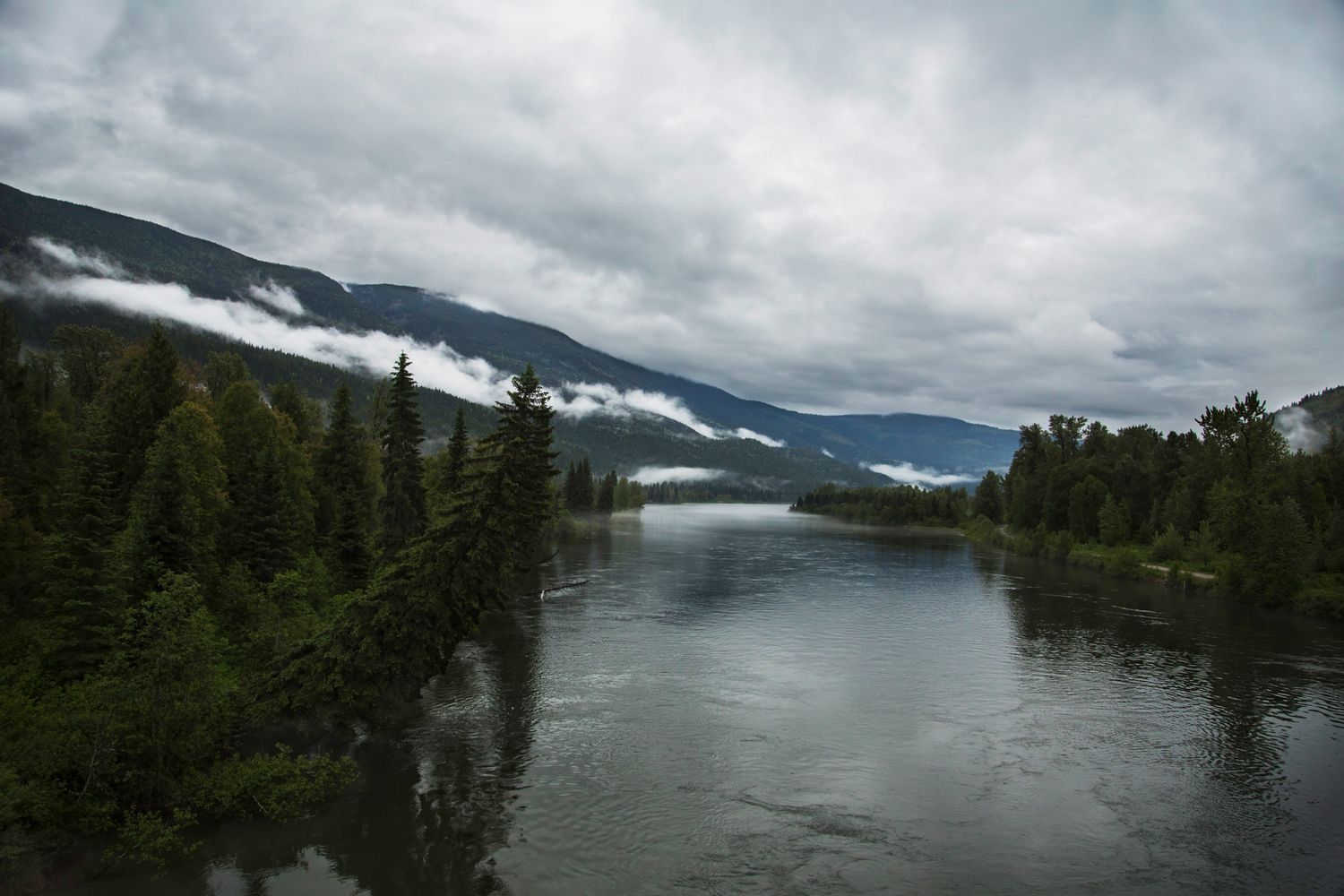 Misty lake surrounded by evergreen forest with snow-capped mountains in distance.