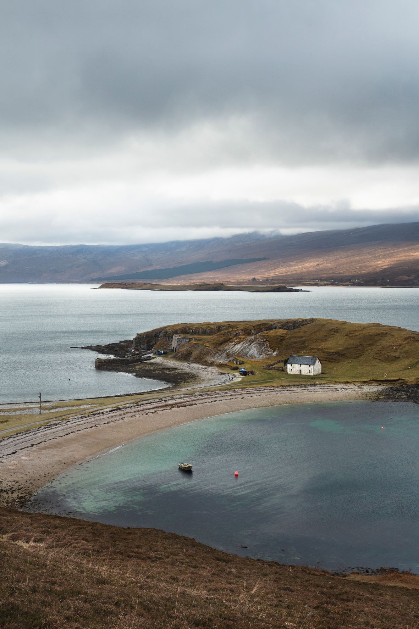 Panoramic coastal view of a remote Scottish bay with white cottage and sandy beach against moody cloudy skies.