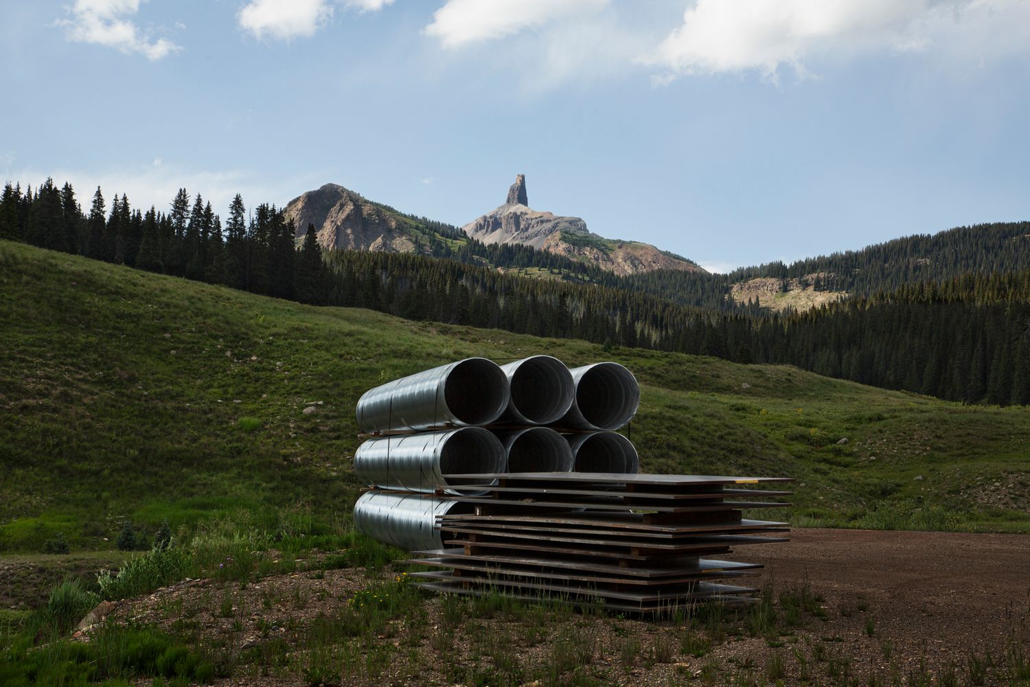 Metal culverts and drainage pipes lie on a grassy mountainside with rugged peaks visible in the background.
