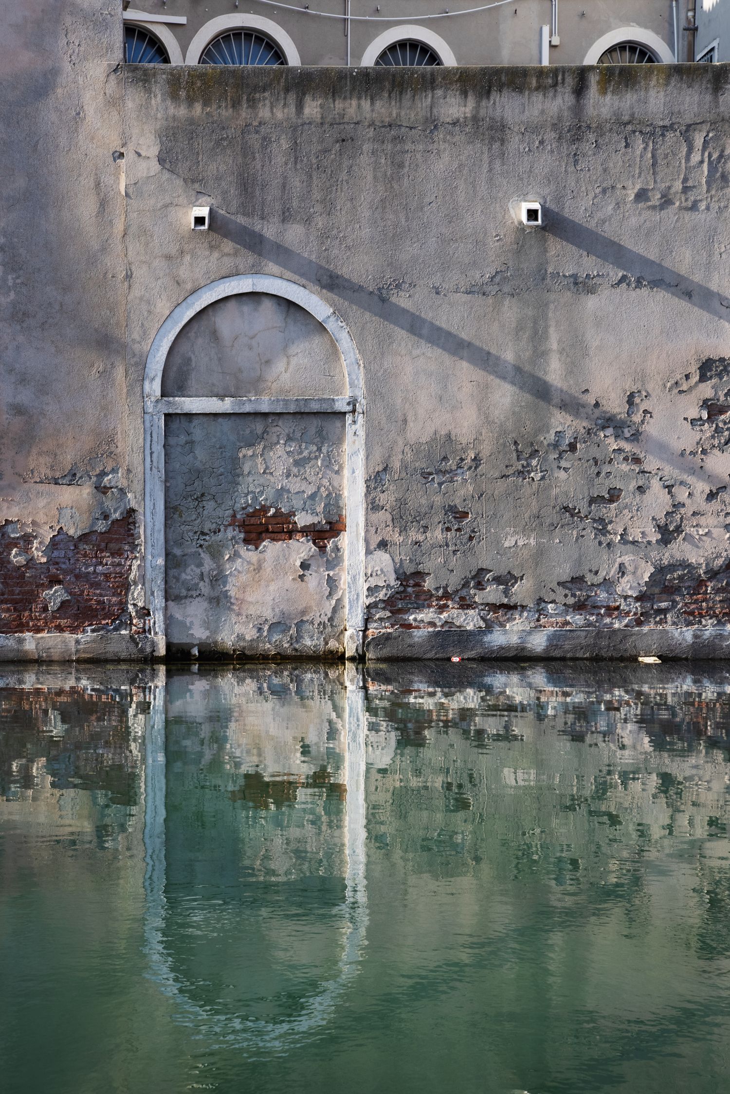 Weathered Venice canal building with arched door reflecting in turquoise green water, aged peeling plaster walls.