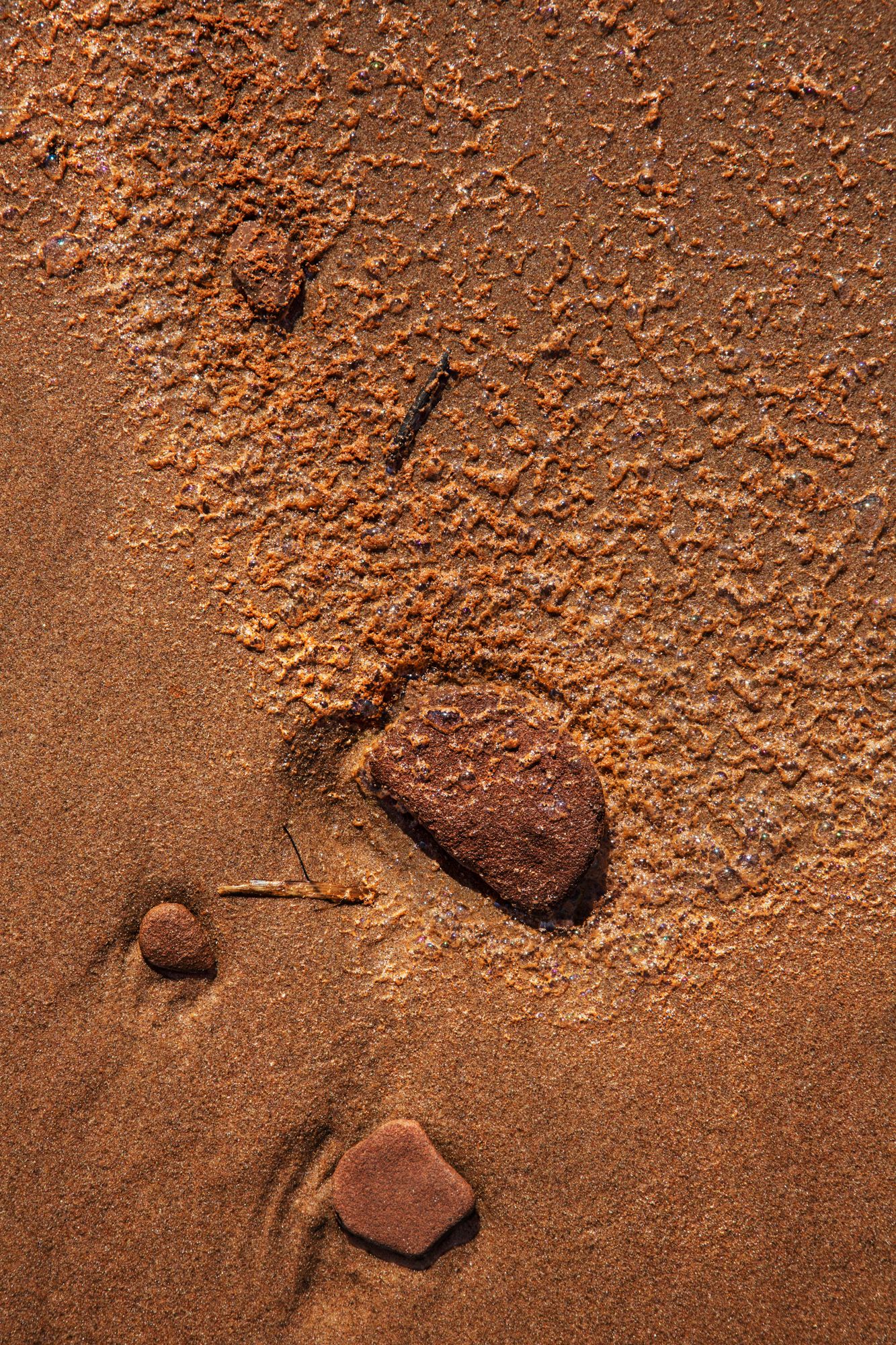 Close-up texture of reddish-brown sand and small rocks on beach terrain.