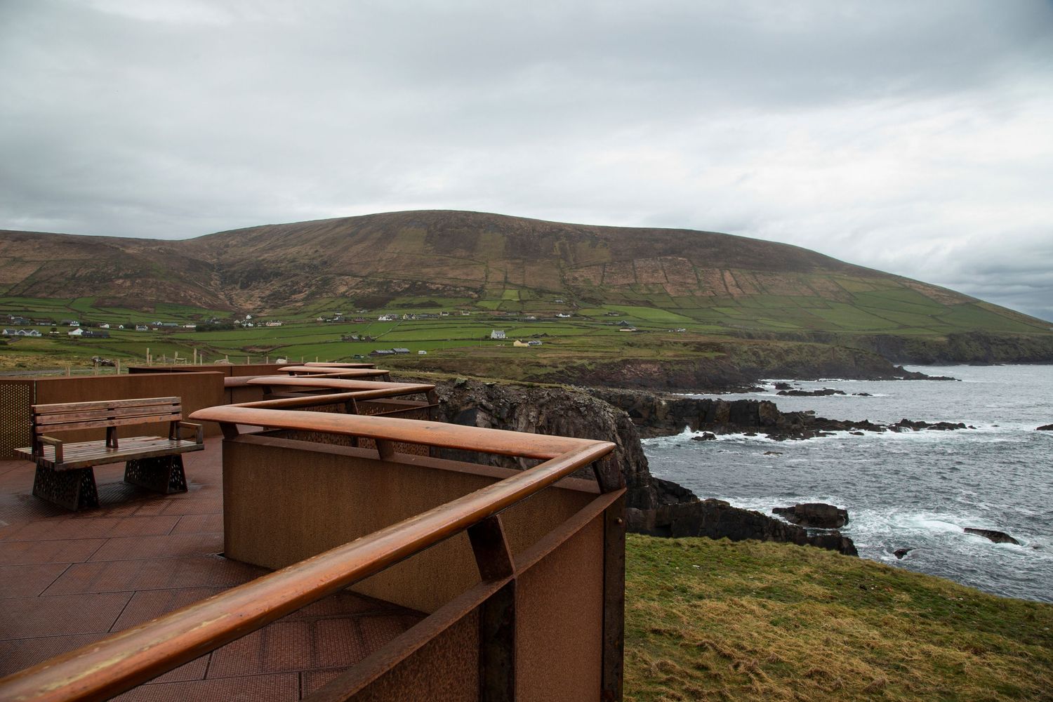 Wooden railing walkway along a rugged coastal cliff with crashing waves and a scenic mountain backdrop.