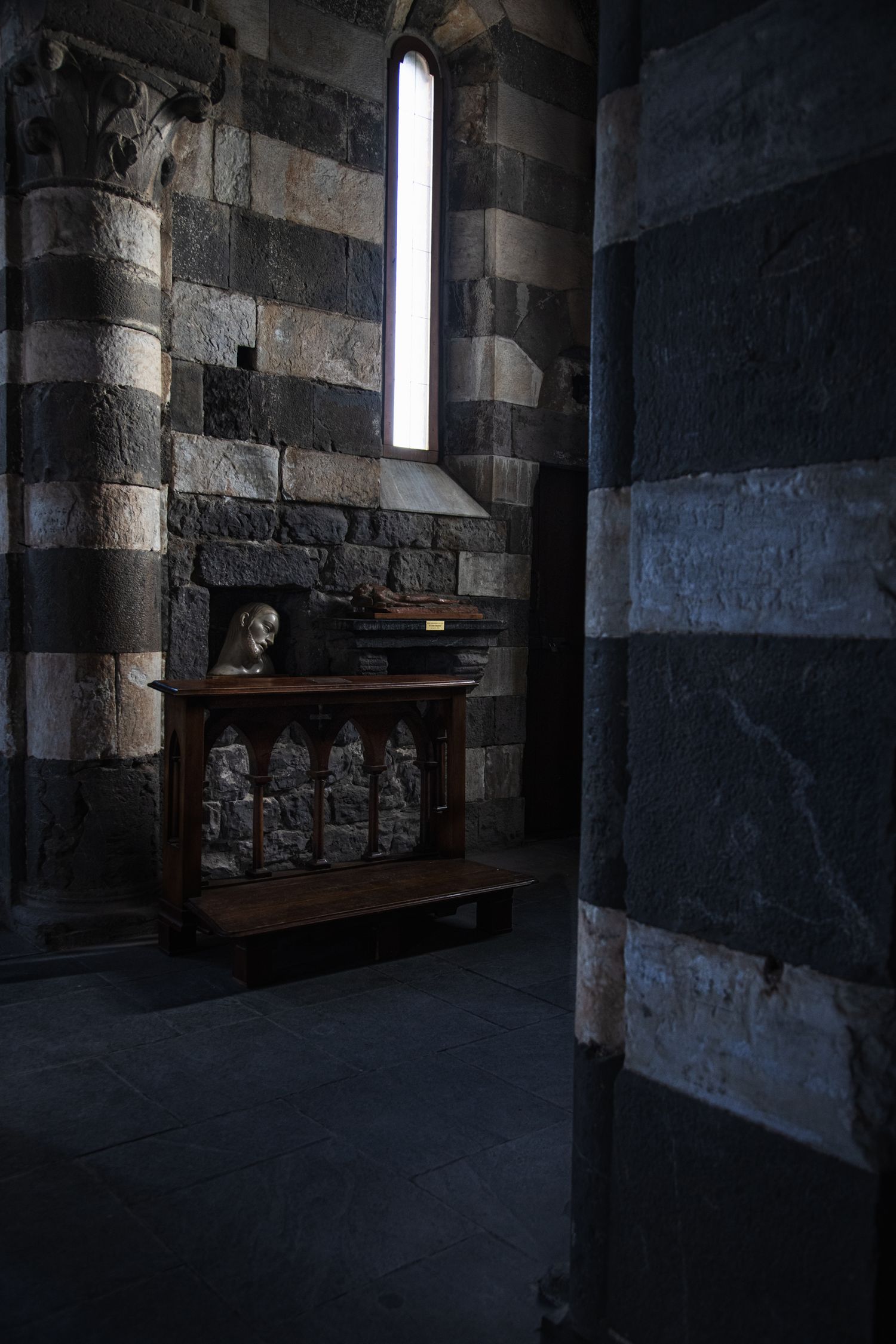 Dark medieval stone church interior with striped walls, wooden altar, and narrow stained glass window.