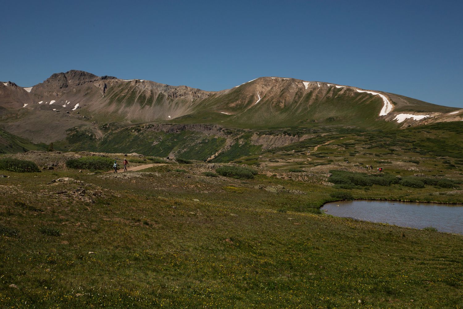 Mountain ridge with patches of snow overlooking green alpine meadow and small lake.