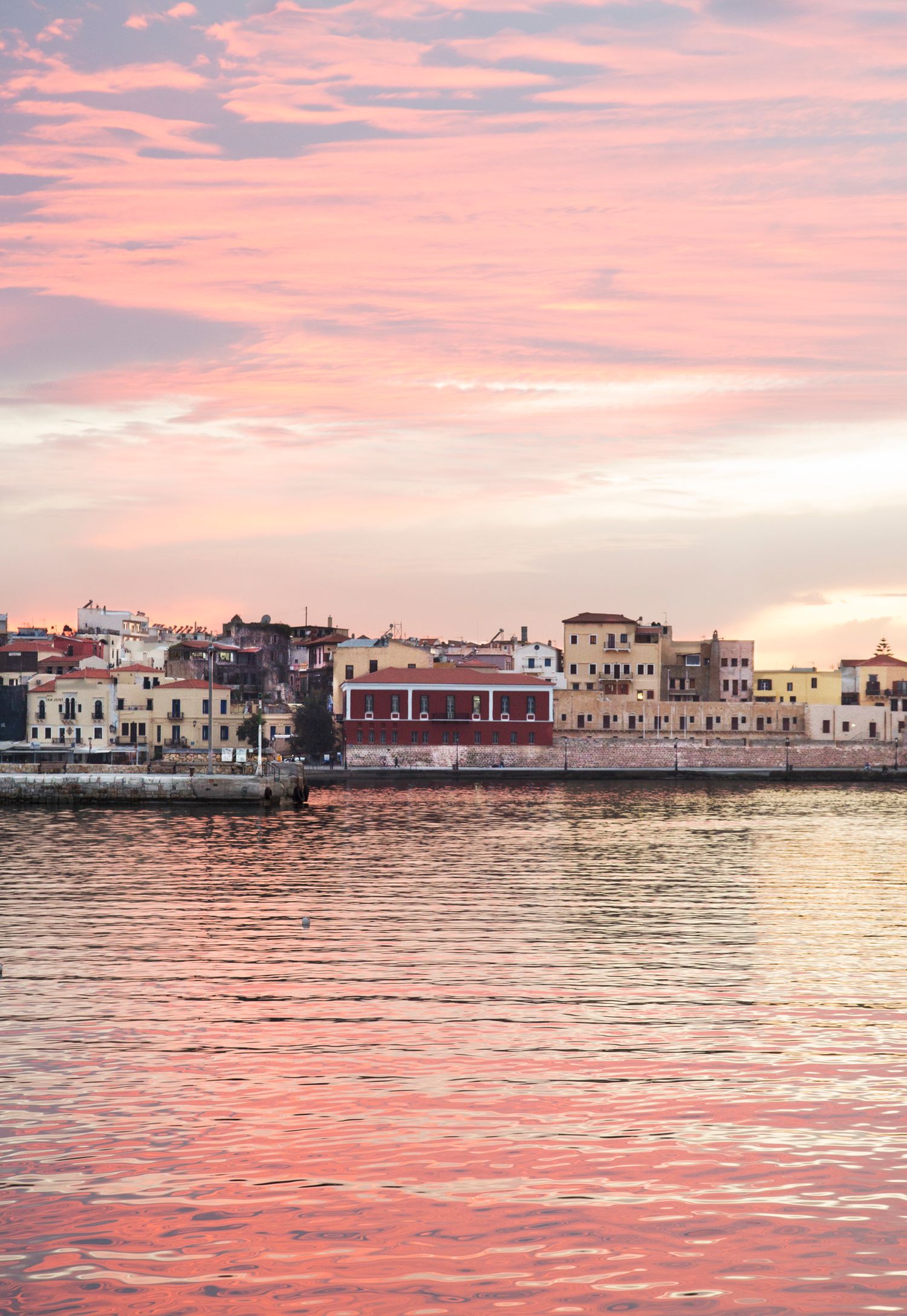 Mediterranean waterfront buildings reflect pink sunset colors on calm waters.