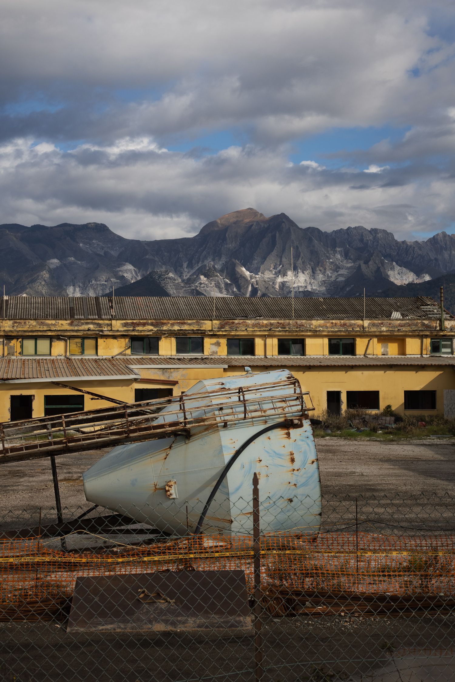 Abandoned industrial facility with rusty tank in foreground, yellow buildings, and dramatic mountain landscape under cloudy sky.