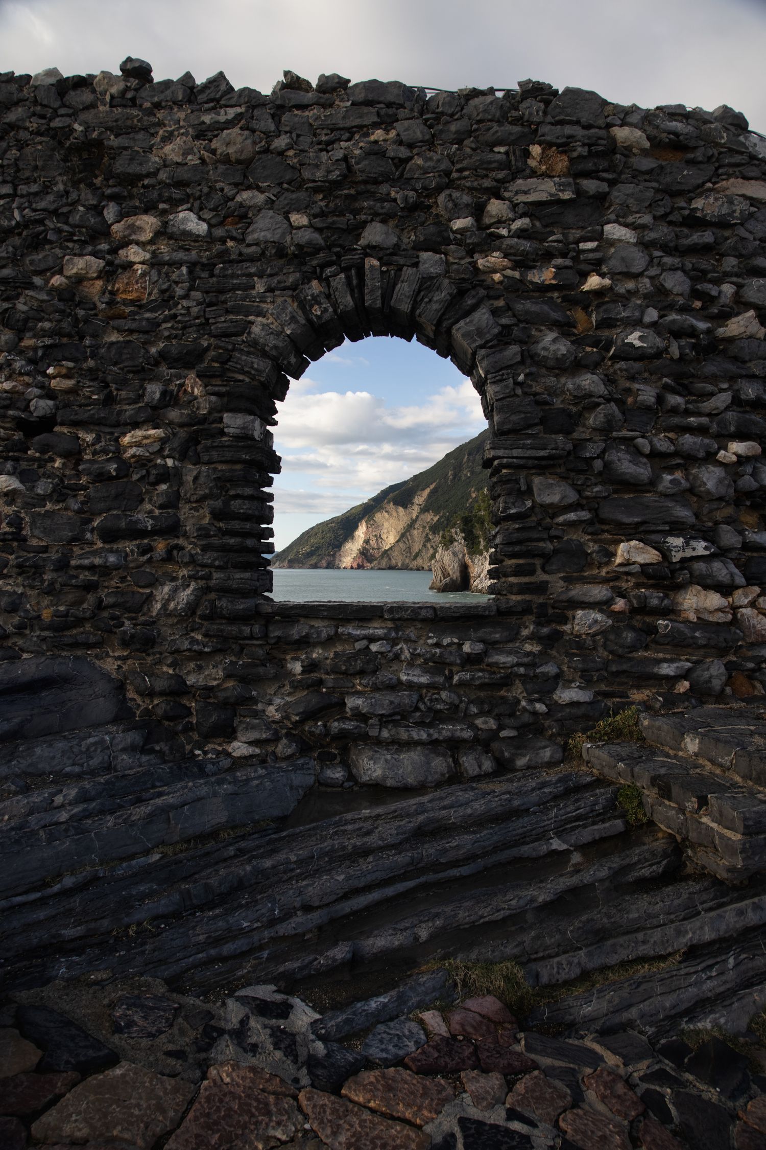 Stone arch window in ancient rocky wall frames a scenic coastal view with cliffs and sea beyond.