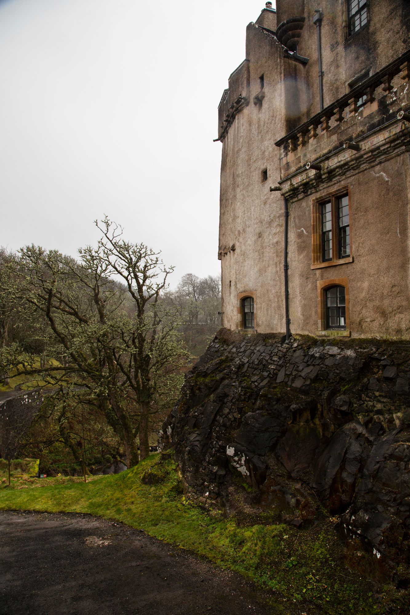 Ancient stone castle walls with dark windows stand against a gloomy sky, surrounded by bare winter trees and green grass.
