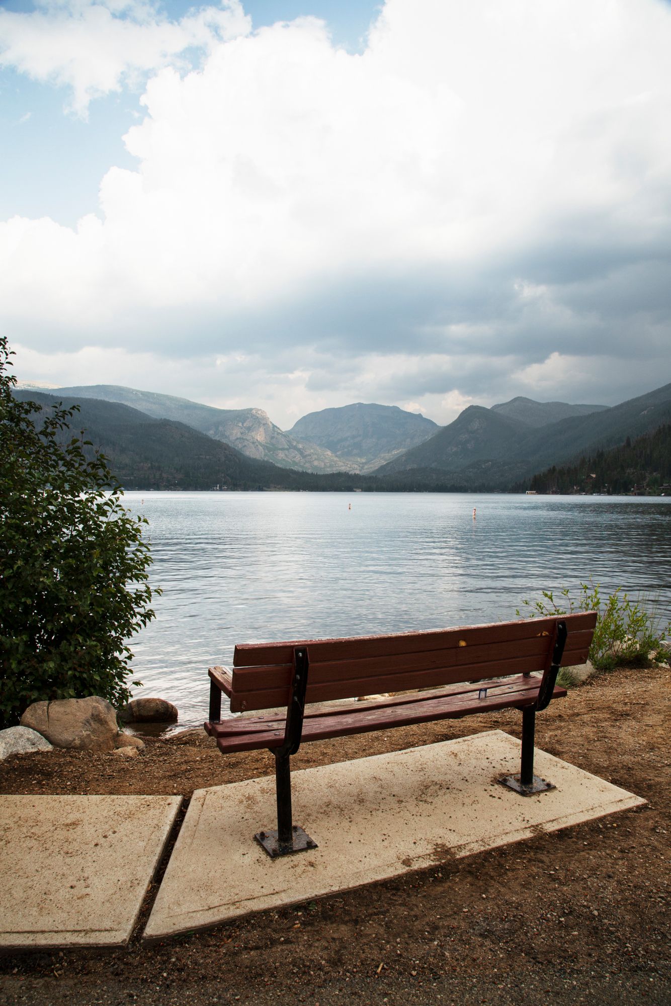 A wooden bench overlooks a serene mountain lake with distant peaks and cloudy skies in a tranquil landscape setting.