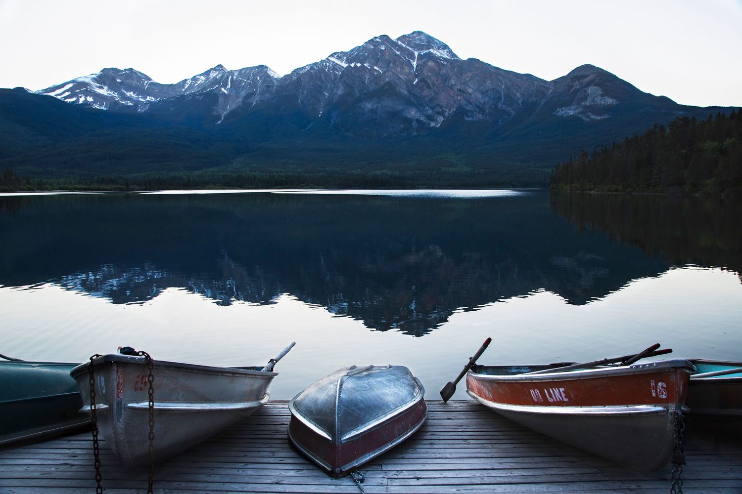 Snow-capped mountain peaks reflect perfectly in a calm lake with wooden boats docked at the shore.