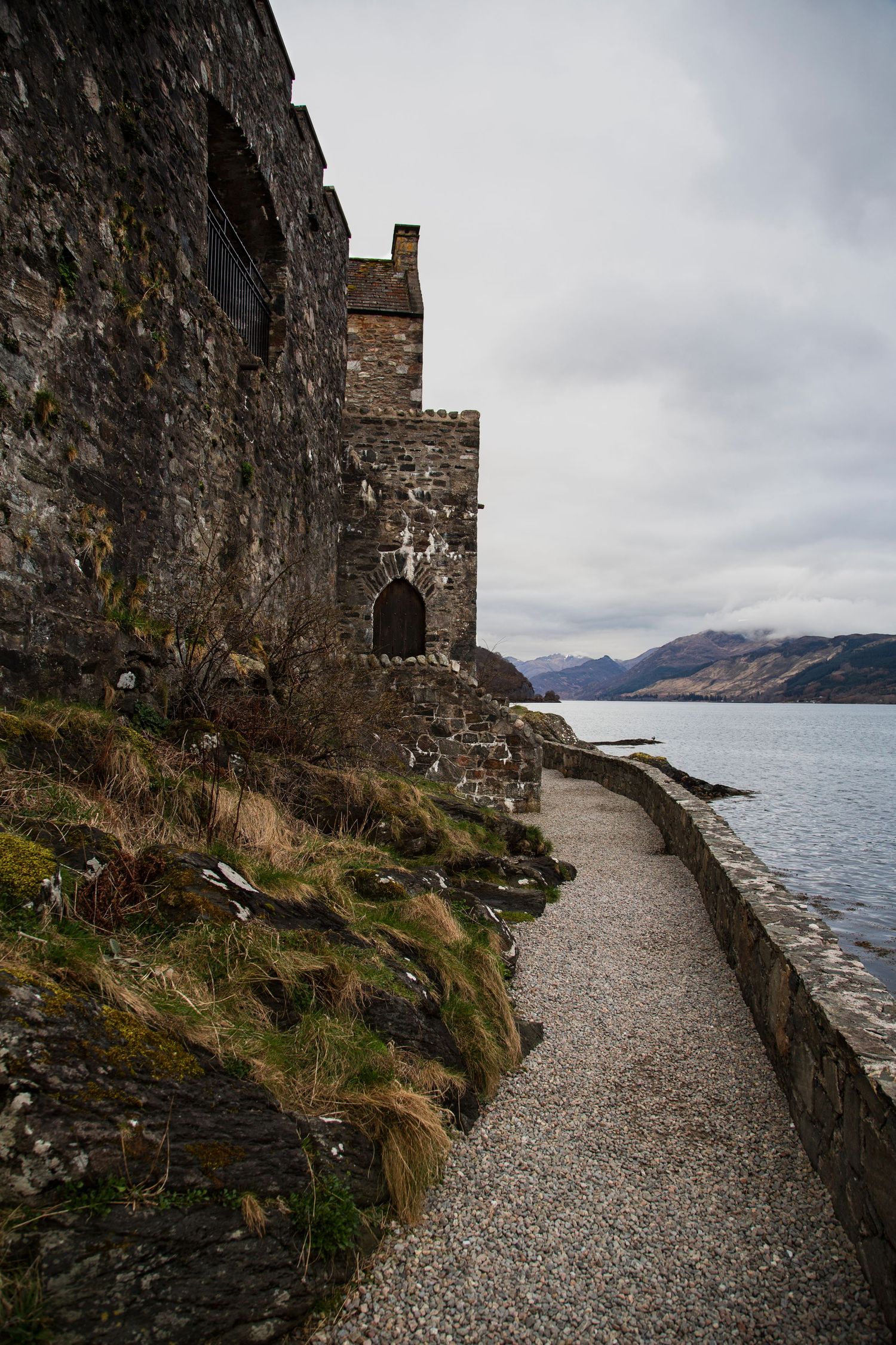 Stone castle pathway along rugged Scottish loch with mountains in distance.