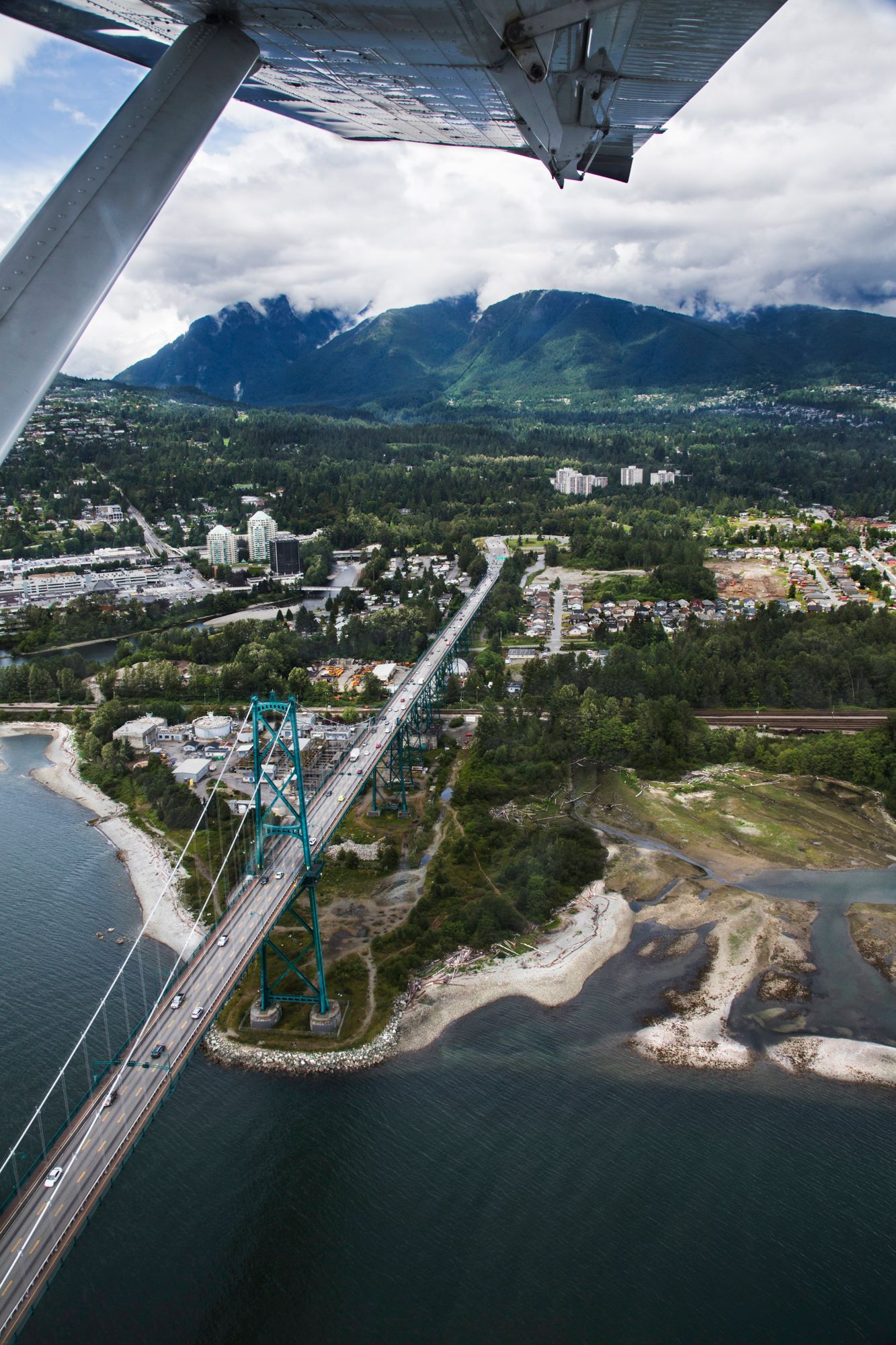 Aerial view of Lions Gate Bridge connecting Vancouver's coastline with snow-capped mountains in background.