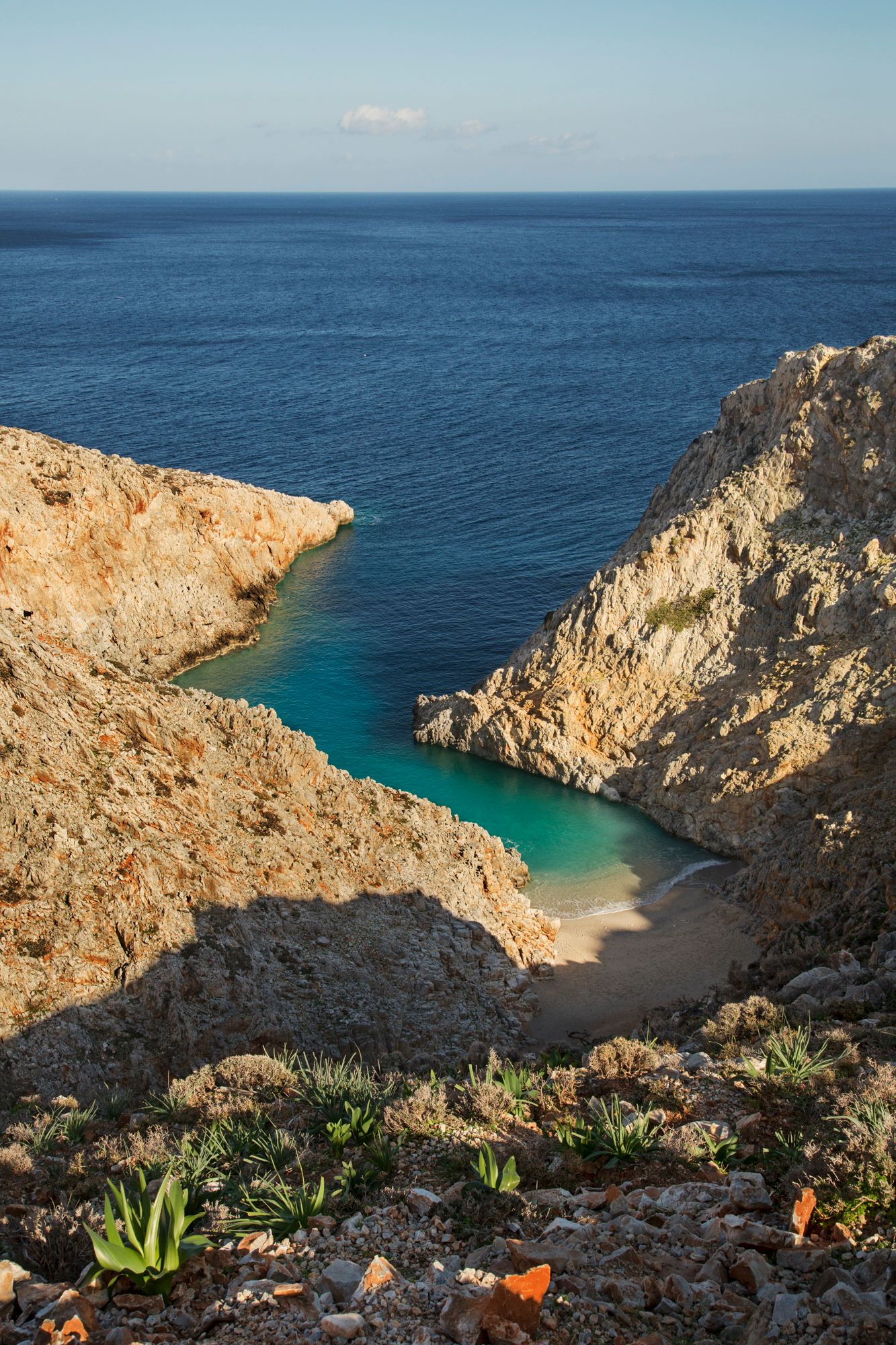Dramatic coastal cliffs frame a secluded turquoise cove with crystal clear waters and rocky outcrops against a blue ocean vista.