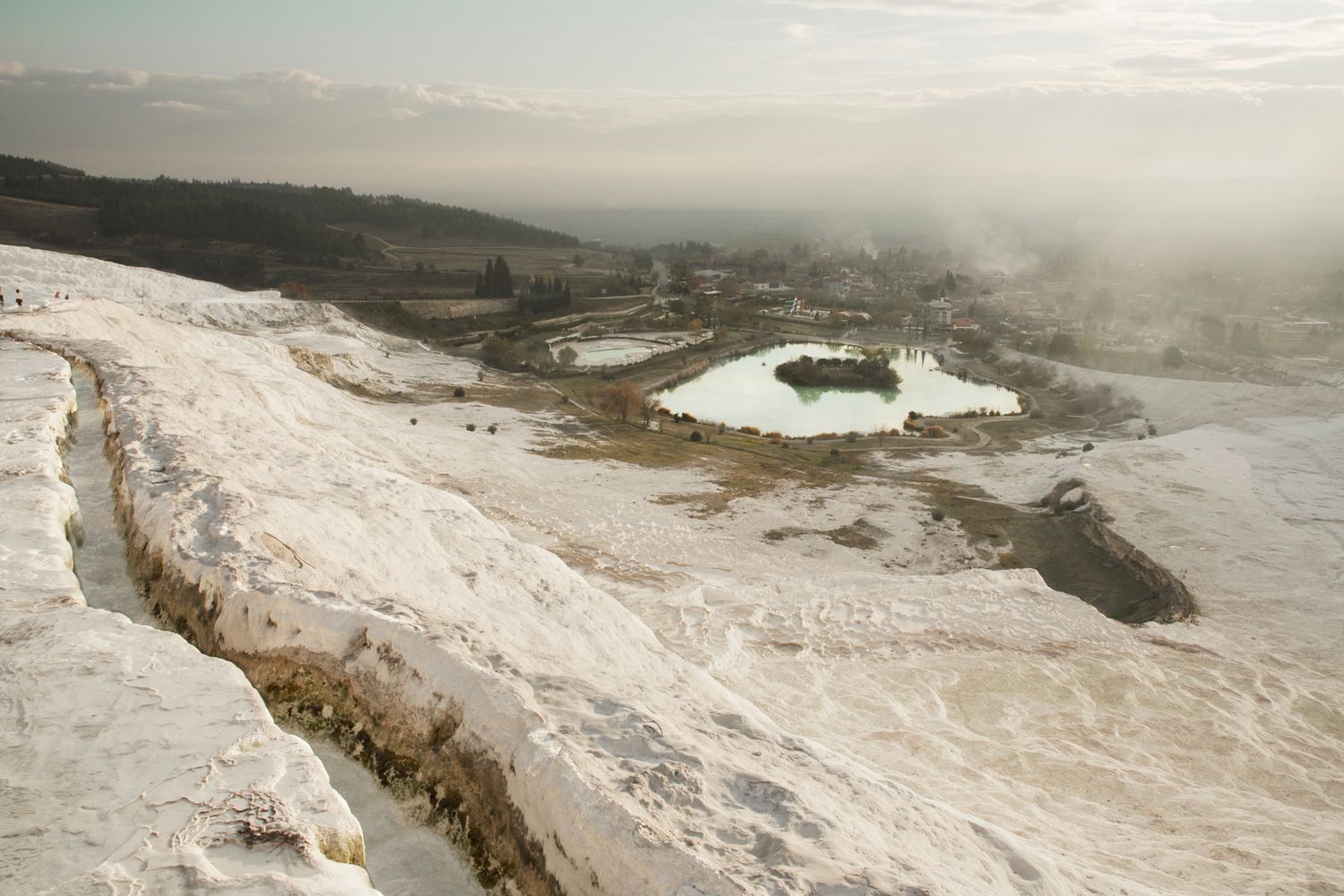 Natural white travertine terraces with thermal pools overlooking misty valley landscape.