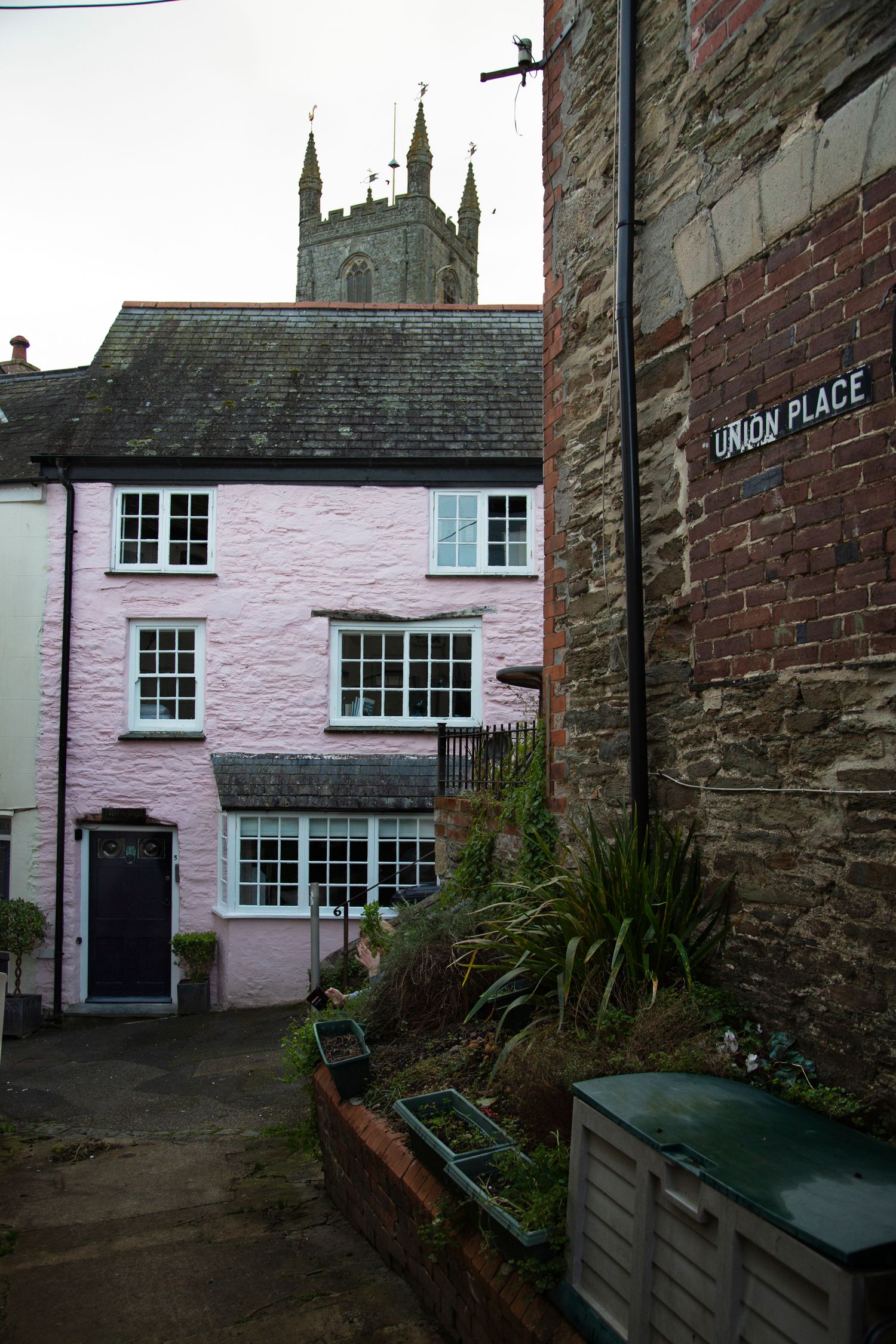 Pink cottage with white windows beneath a Gothic church tower in a narrow alley.