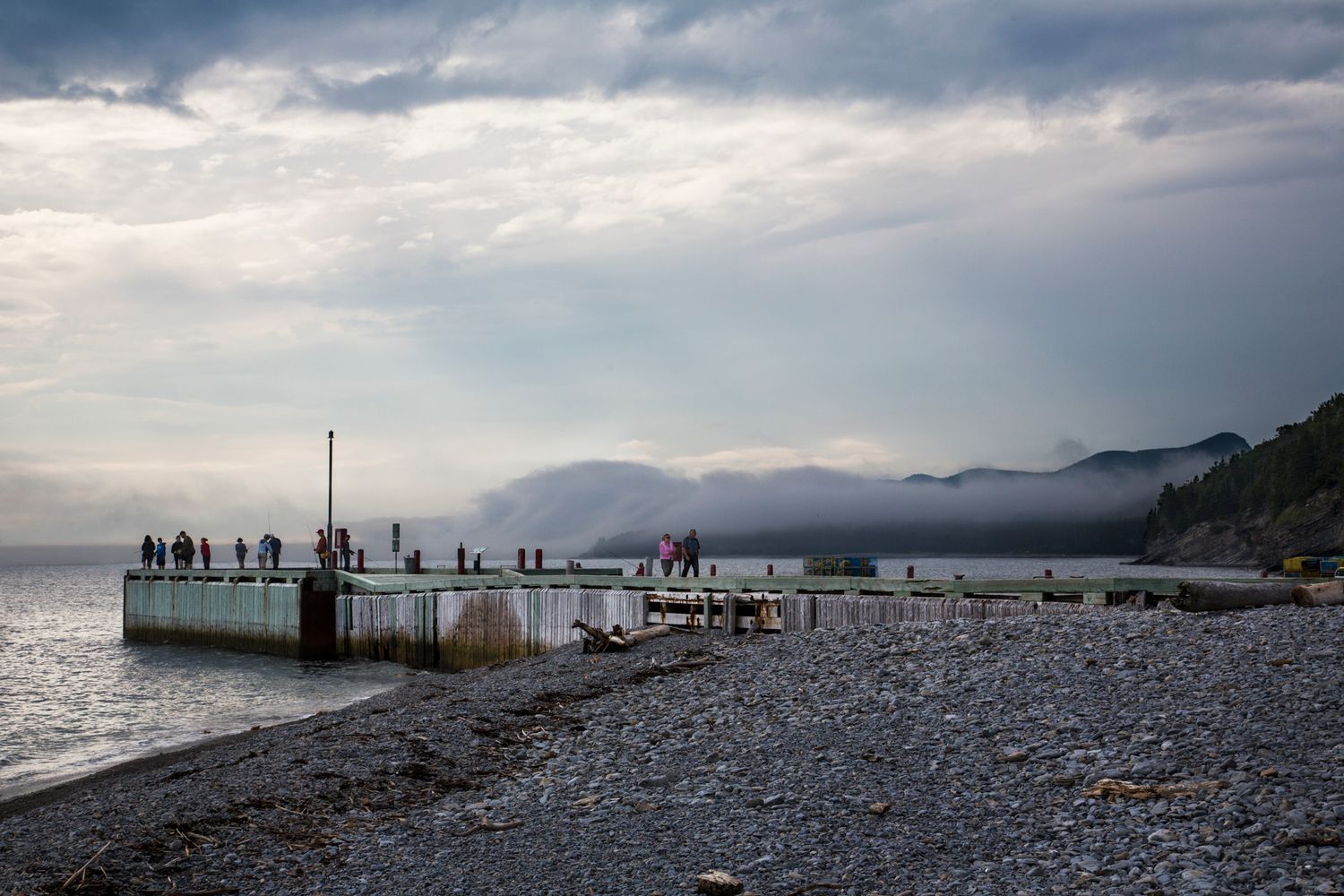 People gathered on rocky beach pier at dusk with mountains in background.
