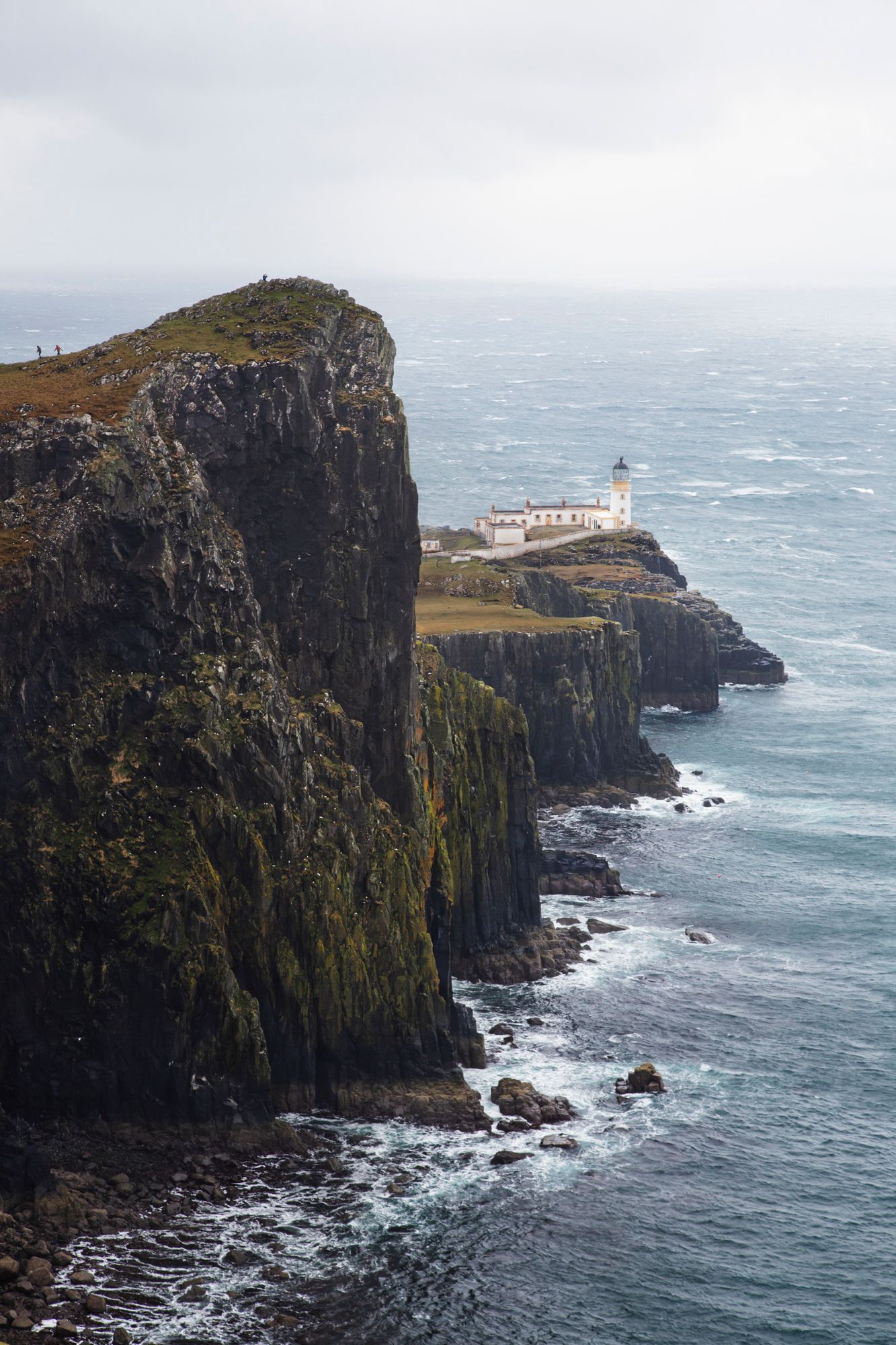 Dramatic coastal cliffs rise above the turbulent ocean with a white lighthouse perched atop at Neist Point, Isle of Skye.