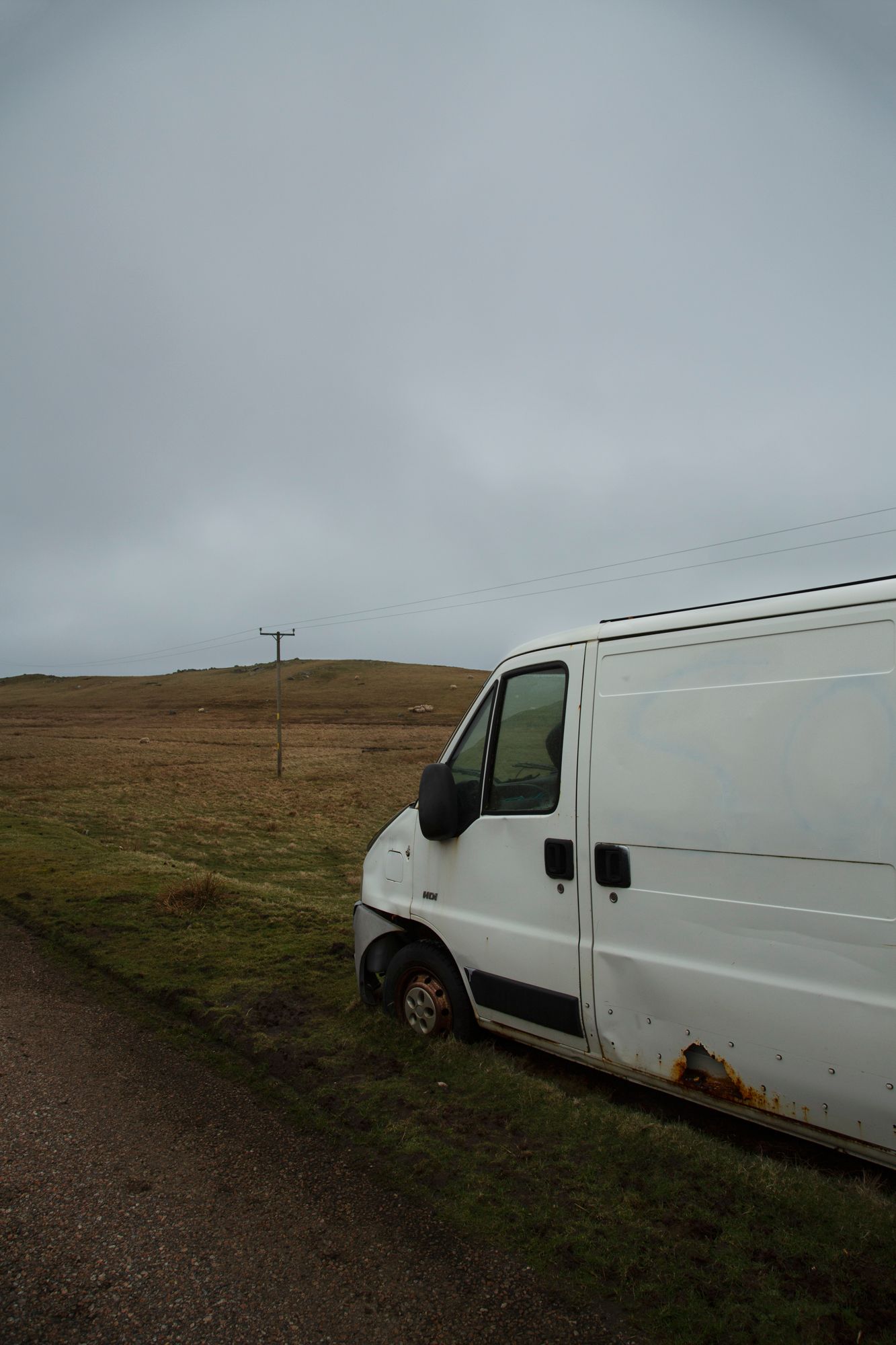 A white cargo van sits parked on a grassy hillside under an overcast sky in a rural landscape.