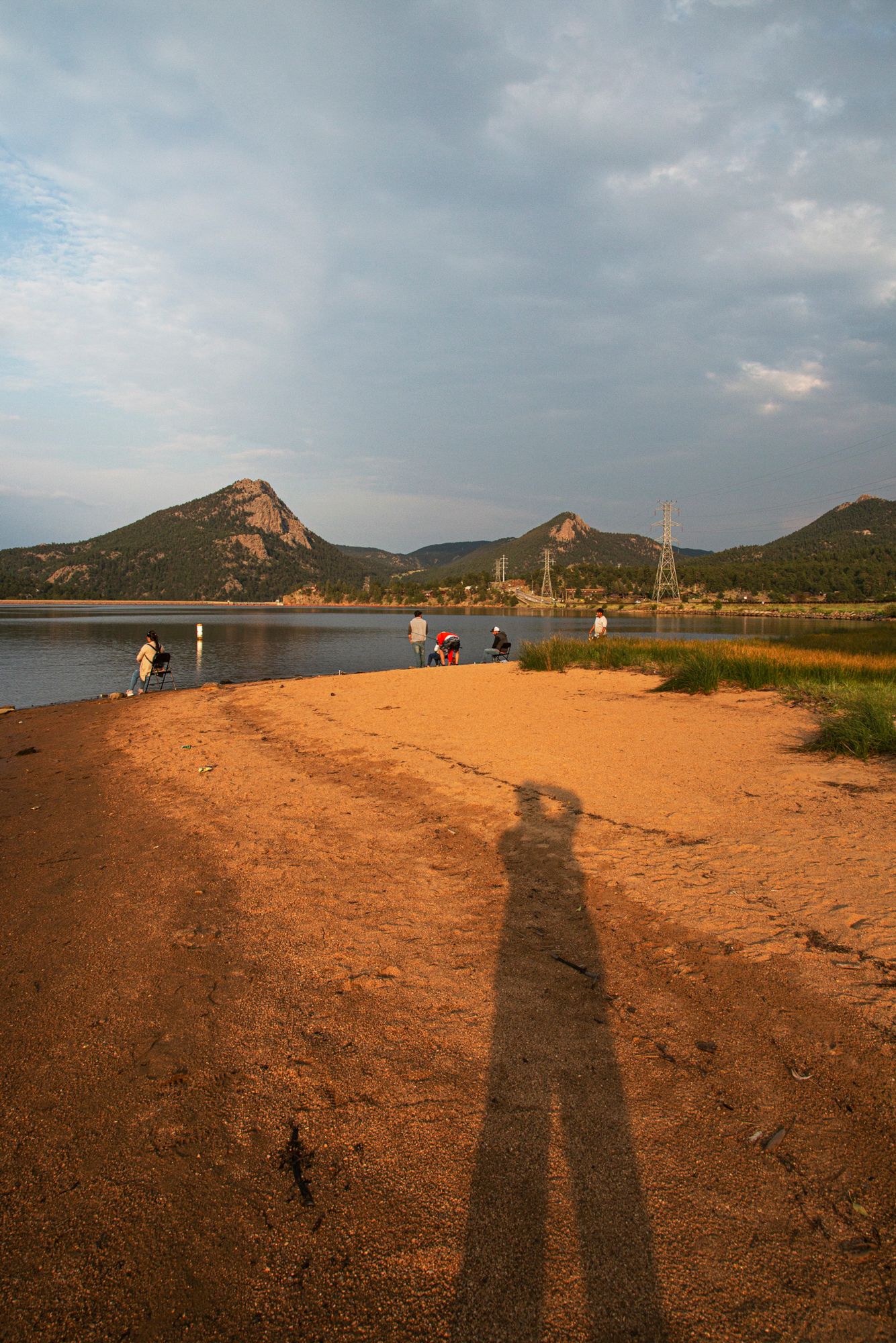 Long shadows stretch across an orange sandy beach with mountains and water visible in the distance during sunset.