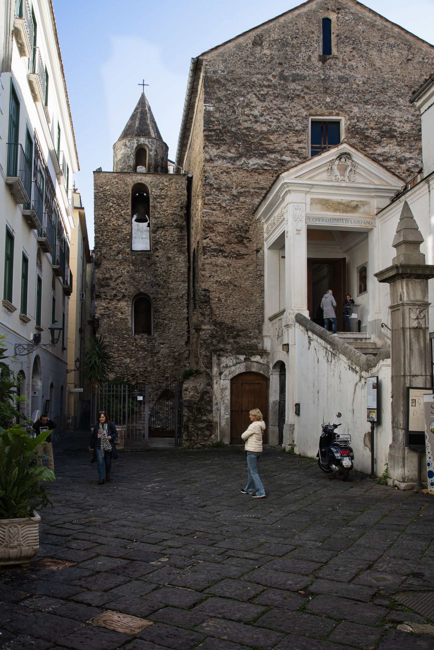Historic Italian piazza with stone church bell tower, cobblestone square, and pedestrians exploring the ancient architecture.