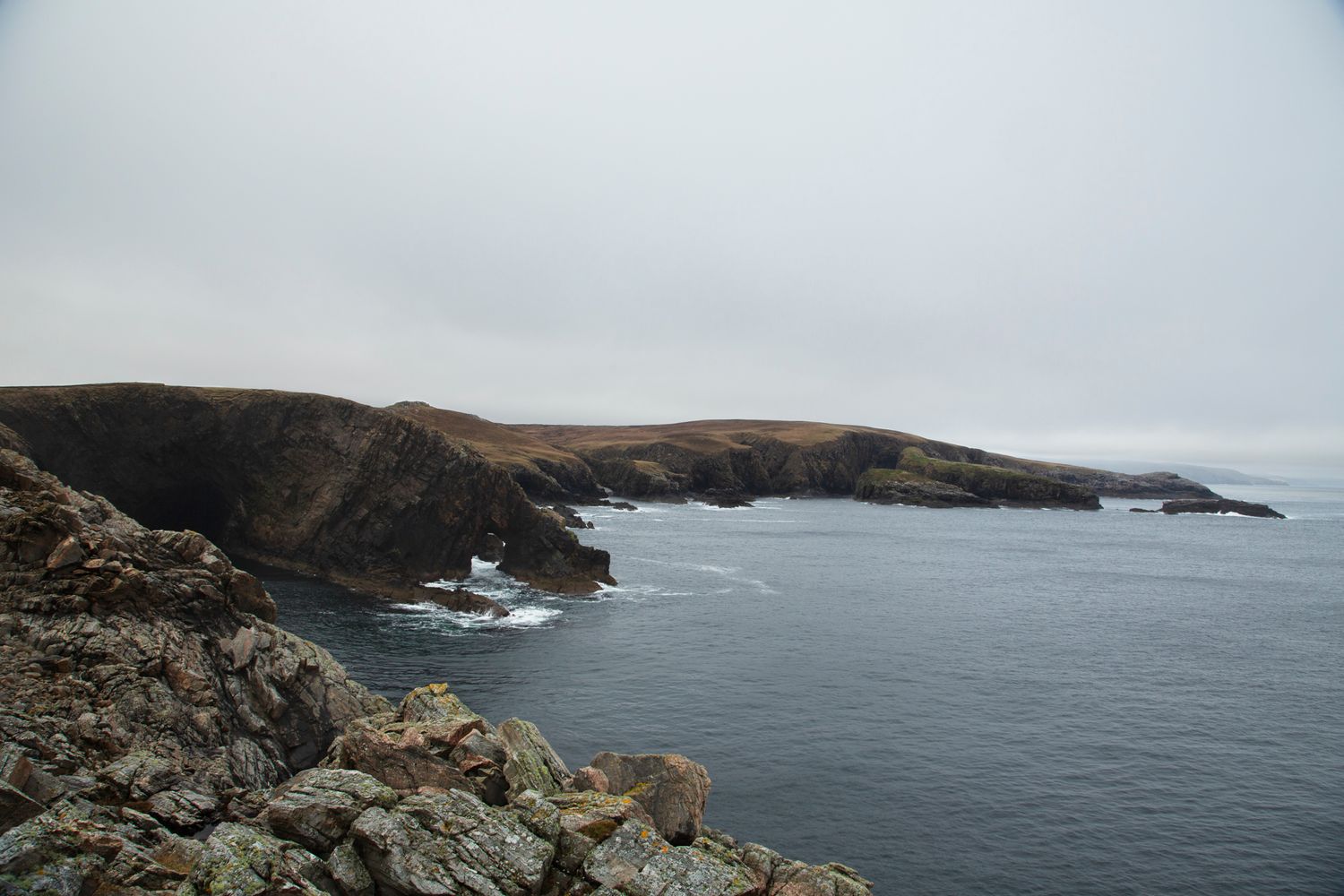 Rocky coastal cliffs overlook a dramatic seascape with waves crashing against the rugged shoreline on a cloudy day.