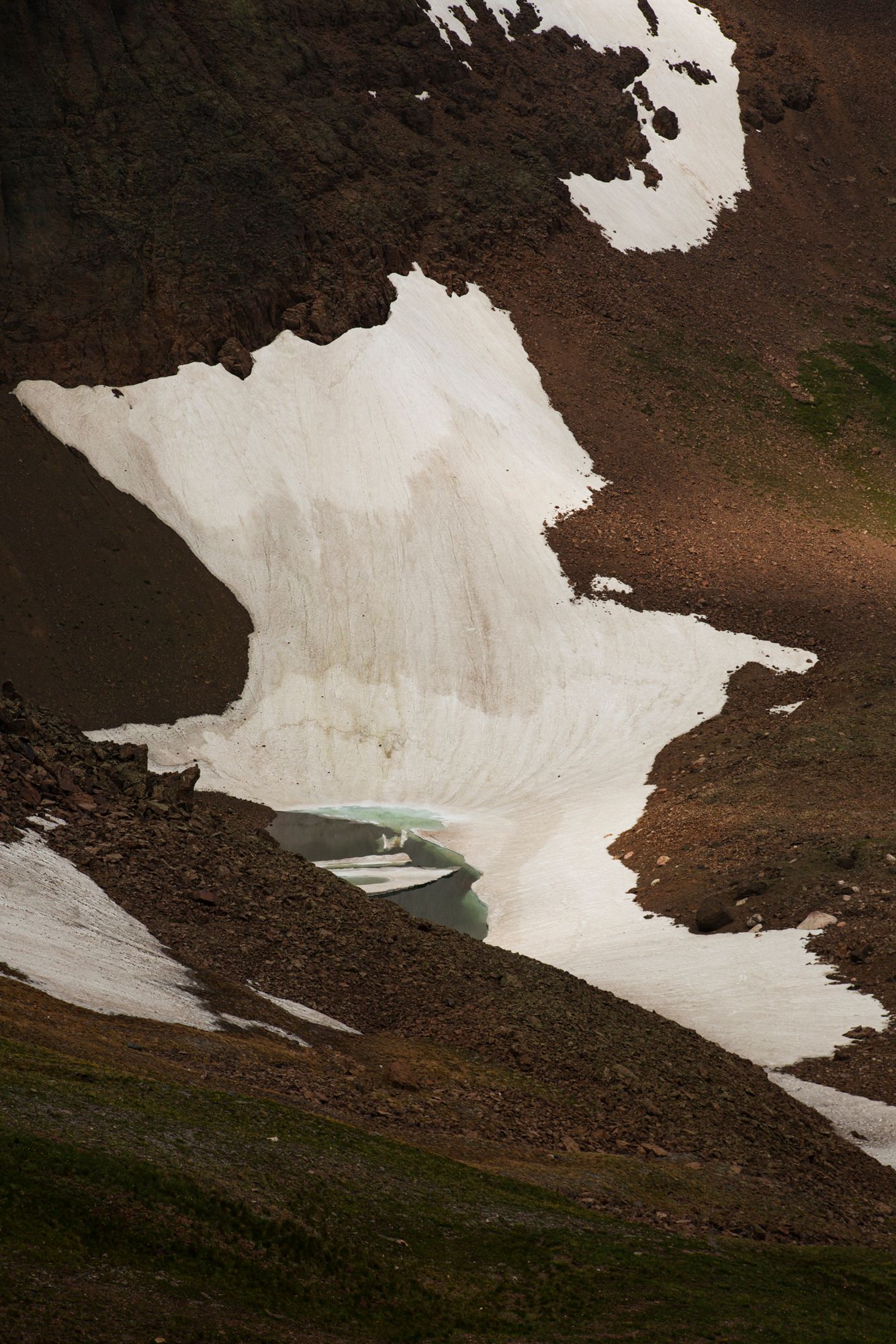 A series of snowy patches on a brown mountainside with small alpine lakes visible in the distance.