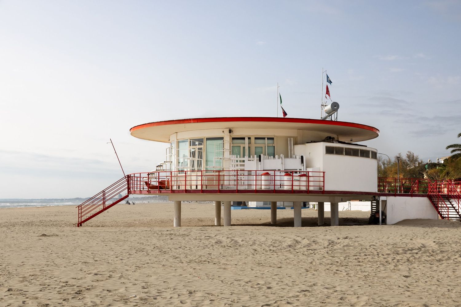 Red and white circular lifeguard station on stilts at a sandy beach with flags flying under a clear sky.