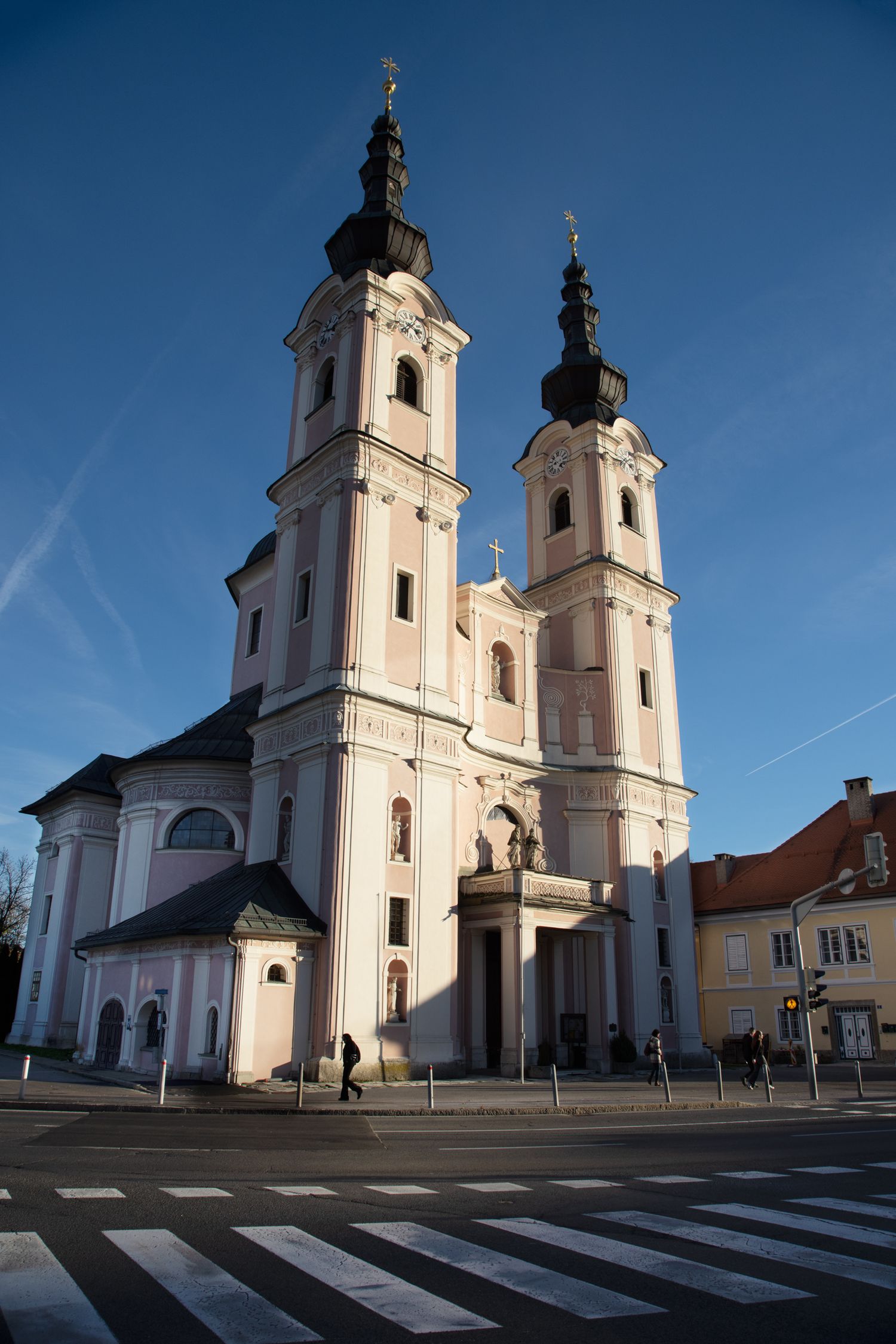 Baroque church with twin black-domed towers and pink facade under a clear blue sky at a street intersection.