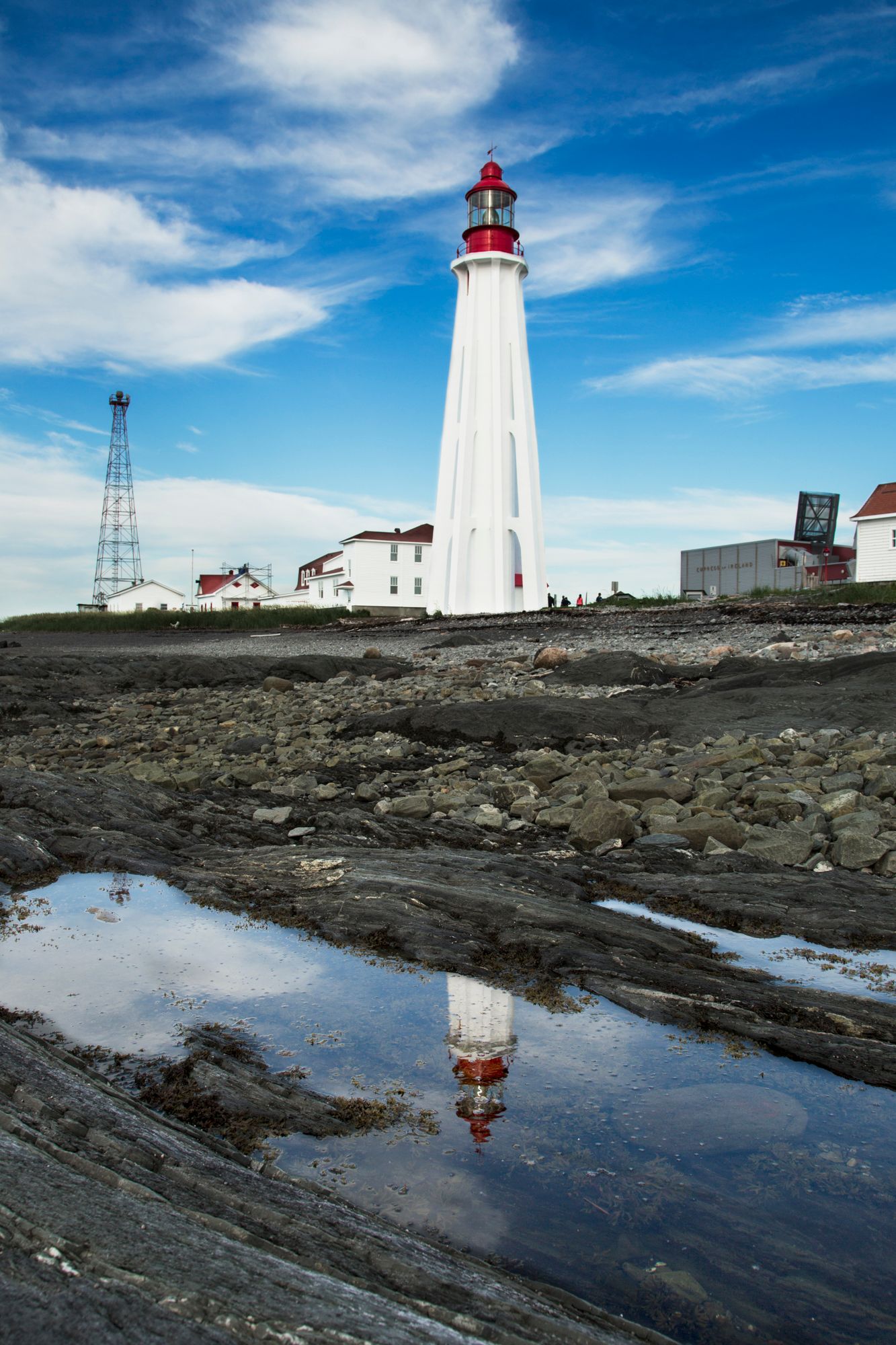 White lighthouse with red top reflected in tide pools on rocky coastal shore under bright blue sky.