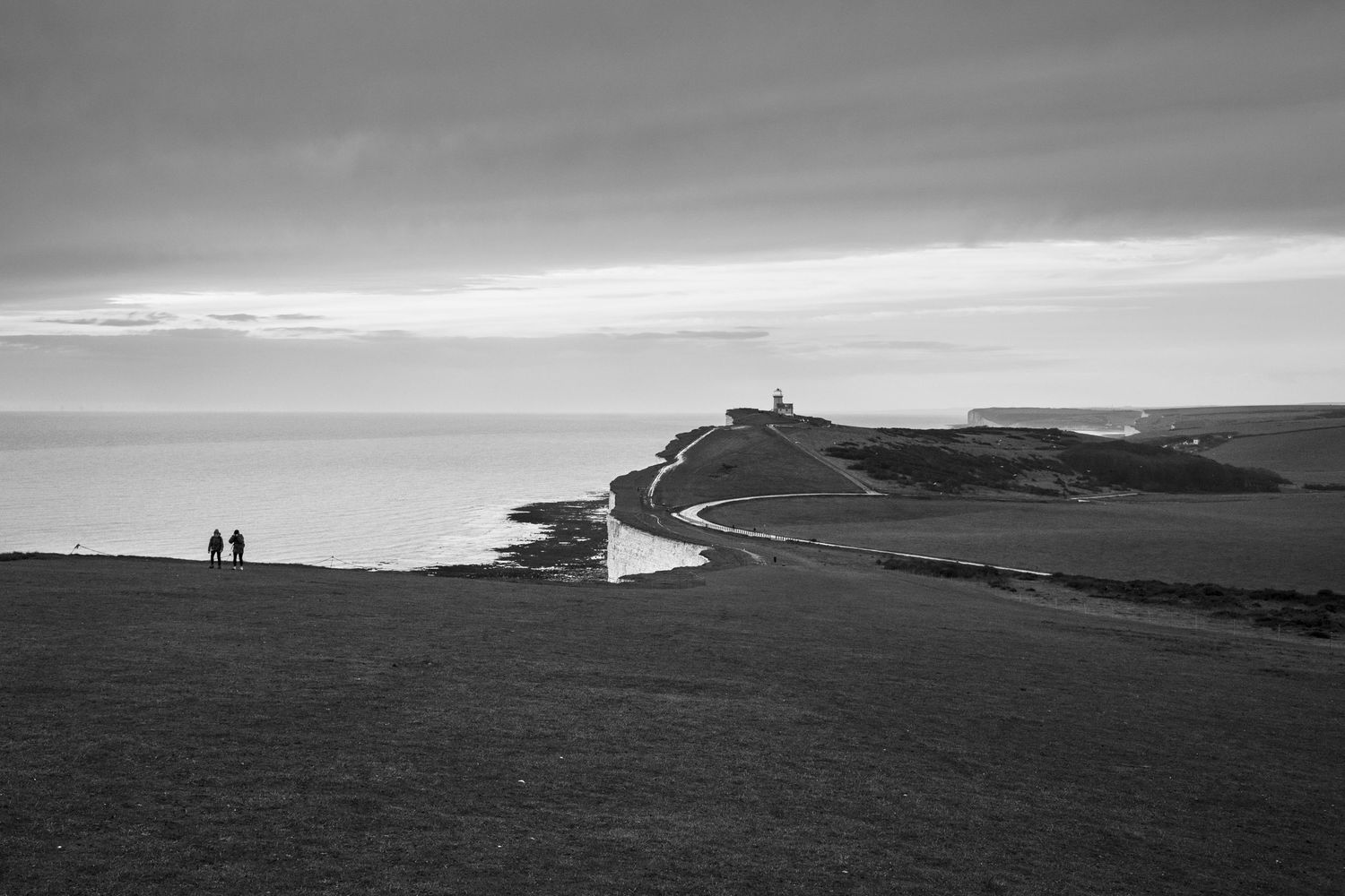 Dramatic black and white coastal landscape with silhouettes against white cliffs and lighthouse in the distance.