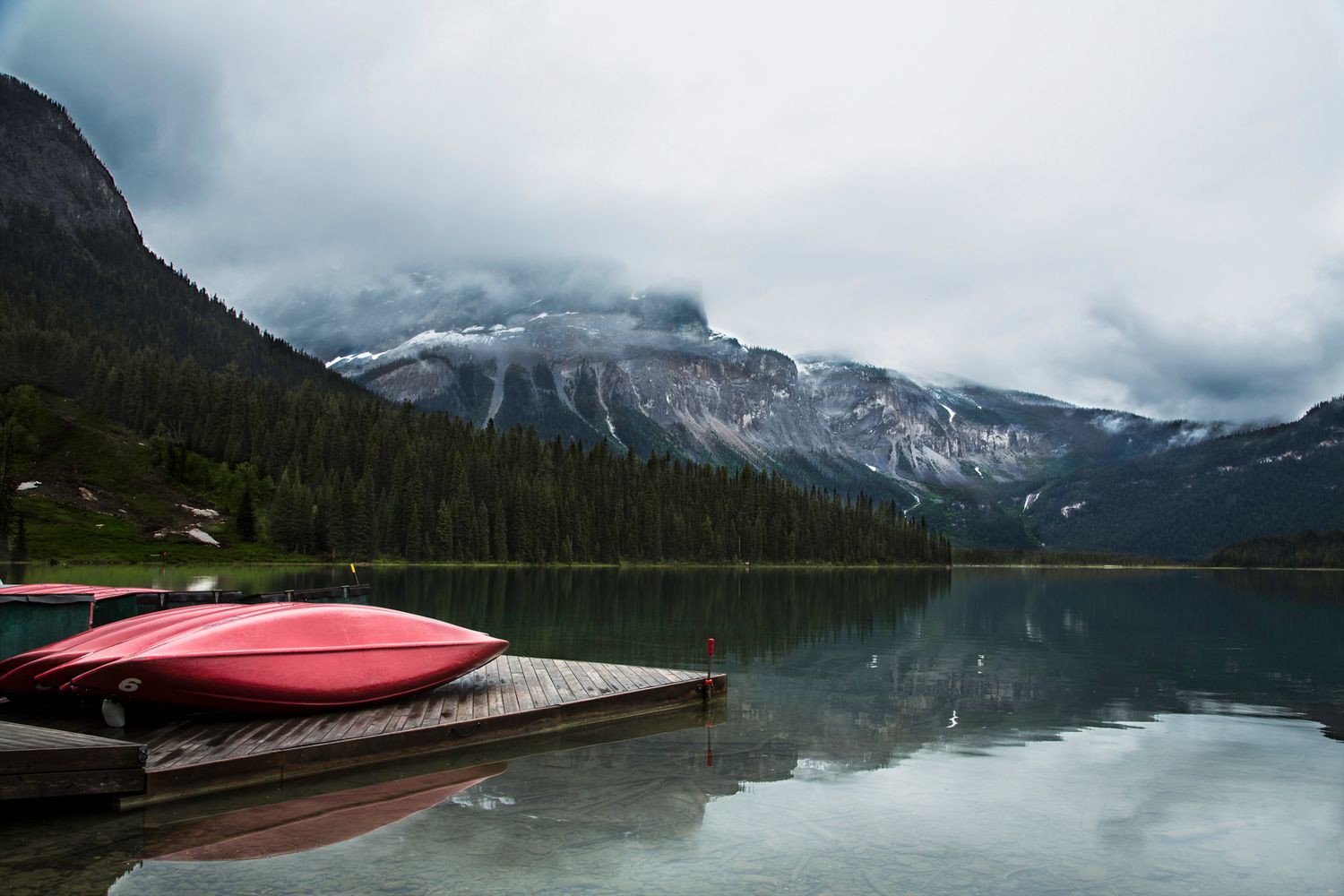 Mountain lake surrounded by evergreen forests with red canoes docked along a wooden pier.