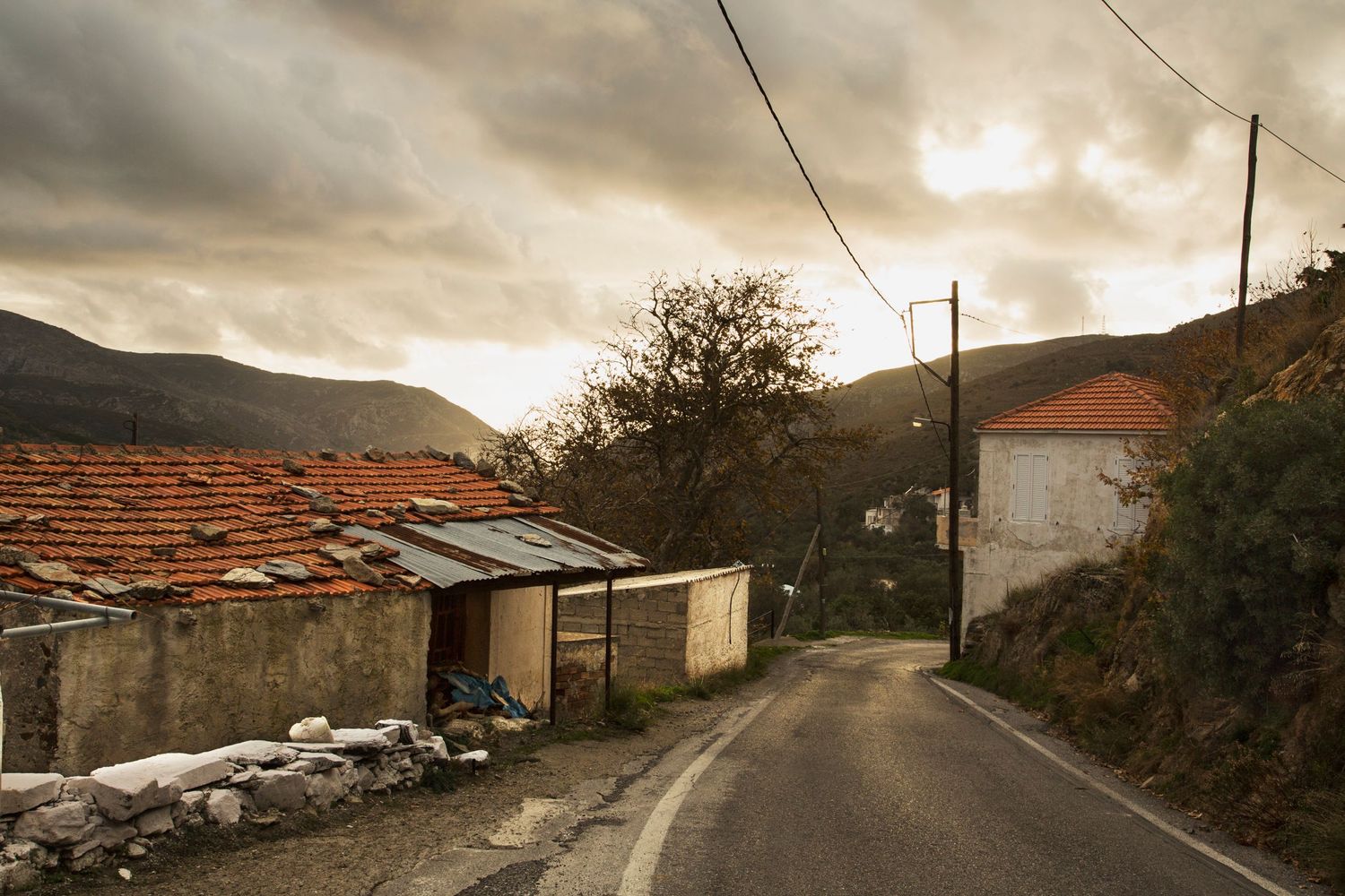 Rural mountain road with dramatic cloudy sky and stone buildings along winding path through rugged terrain.
