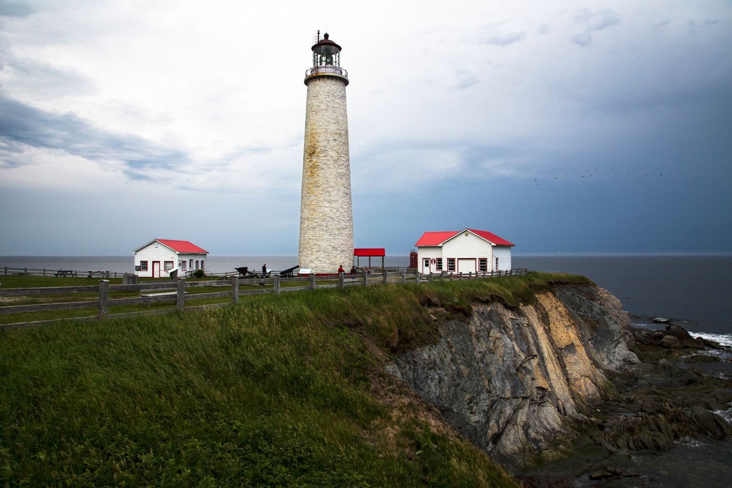 Historic lighthouse with red-roofed buildings perched on rocky coastal cliffs.