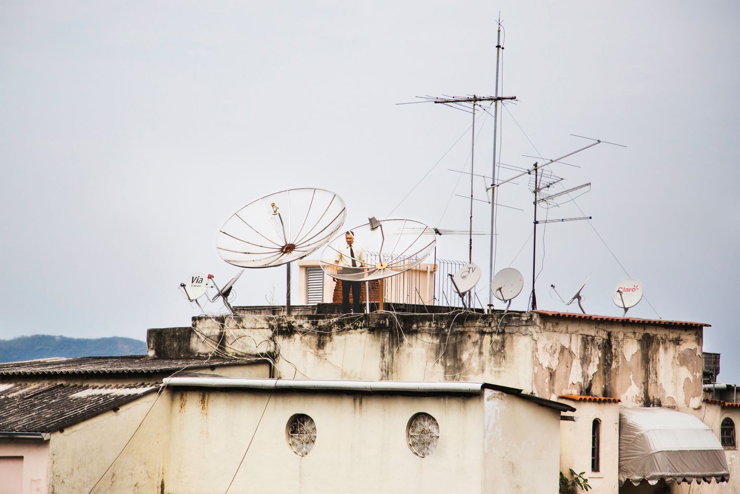 Row of satellite dishes and antennas on building rooftops against a cloudy sky.