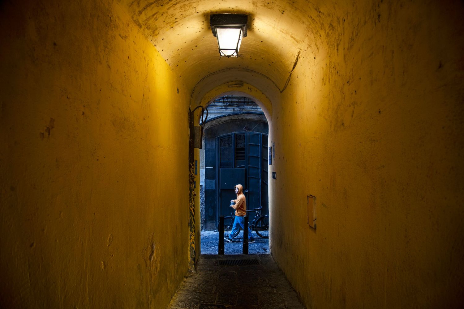 Person silhouetted in a glowing yellow arched tunnel passageway at night, lit by a vintage wall lantern.